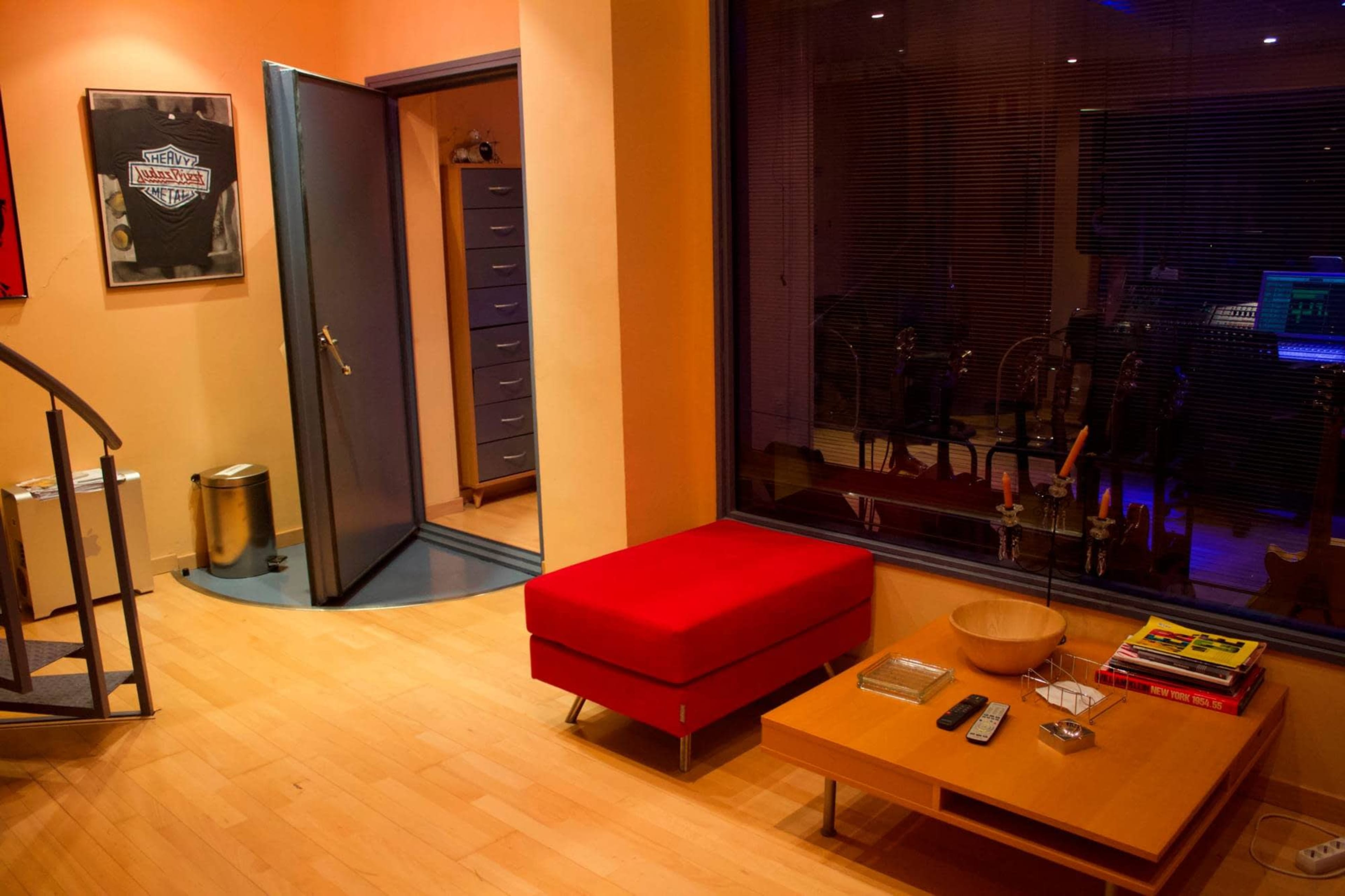 A modern room with a spiral staircase, a red sofa, a wooden coffee table with books and a remote, and a large window revealing an indoor space beyond.