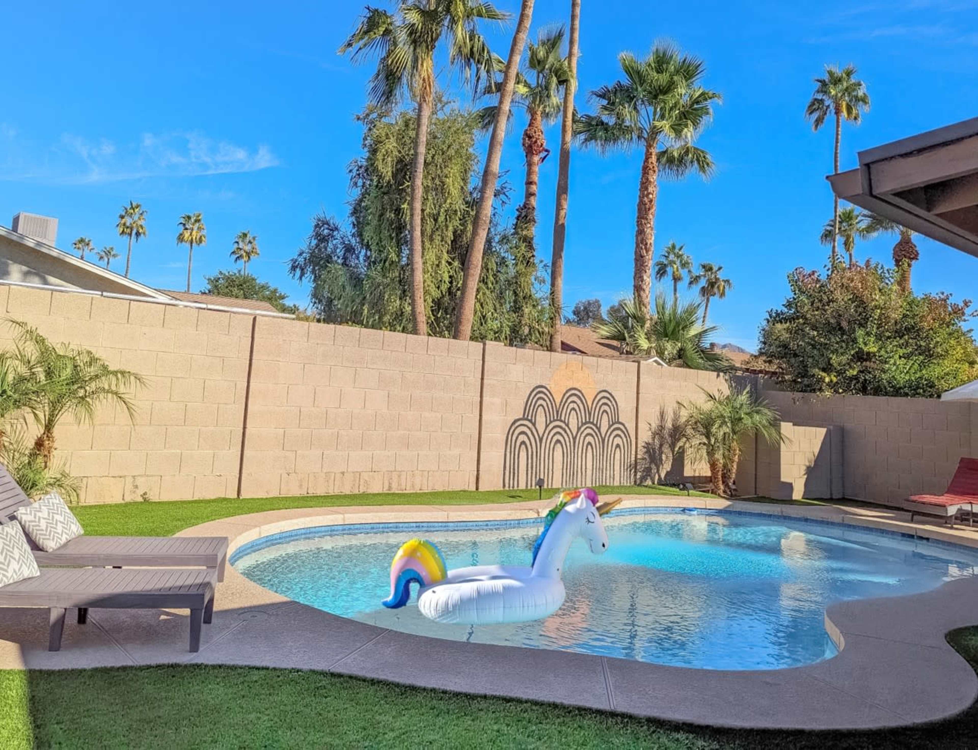 A colorful unicorn float is positioned in a sparkling blue pool surrounded by palm trees and a beige wall in a sunny backyard.