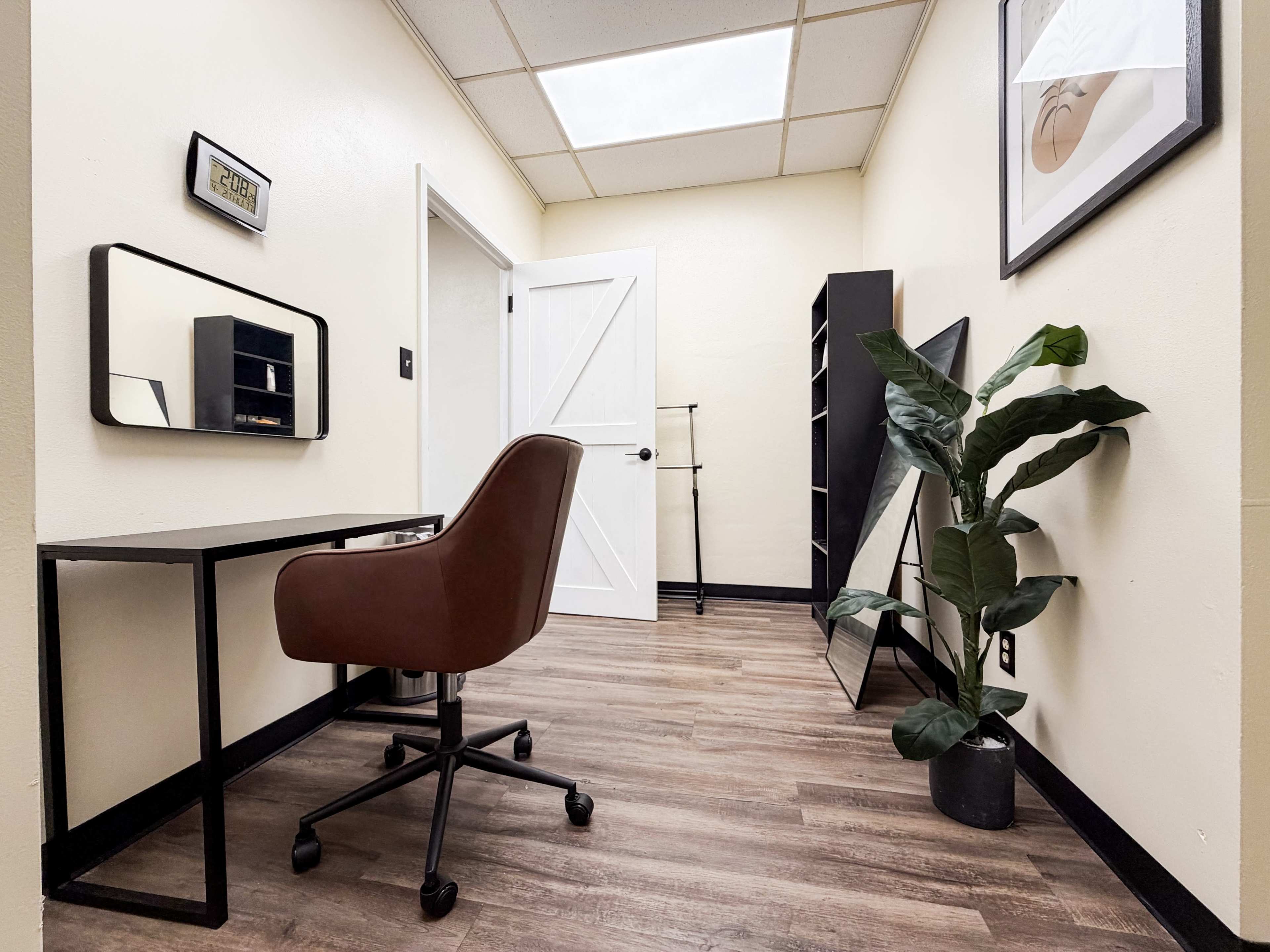 The image shows a minimalist office space featuring a brown rolling chair, a desk with a mirror, and a plant in a pot, with a large mirror and white door in the background.