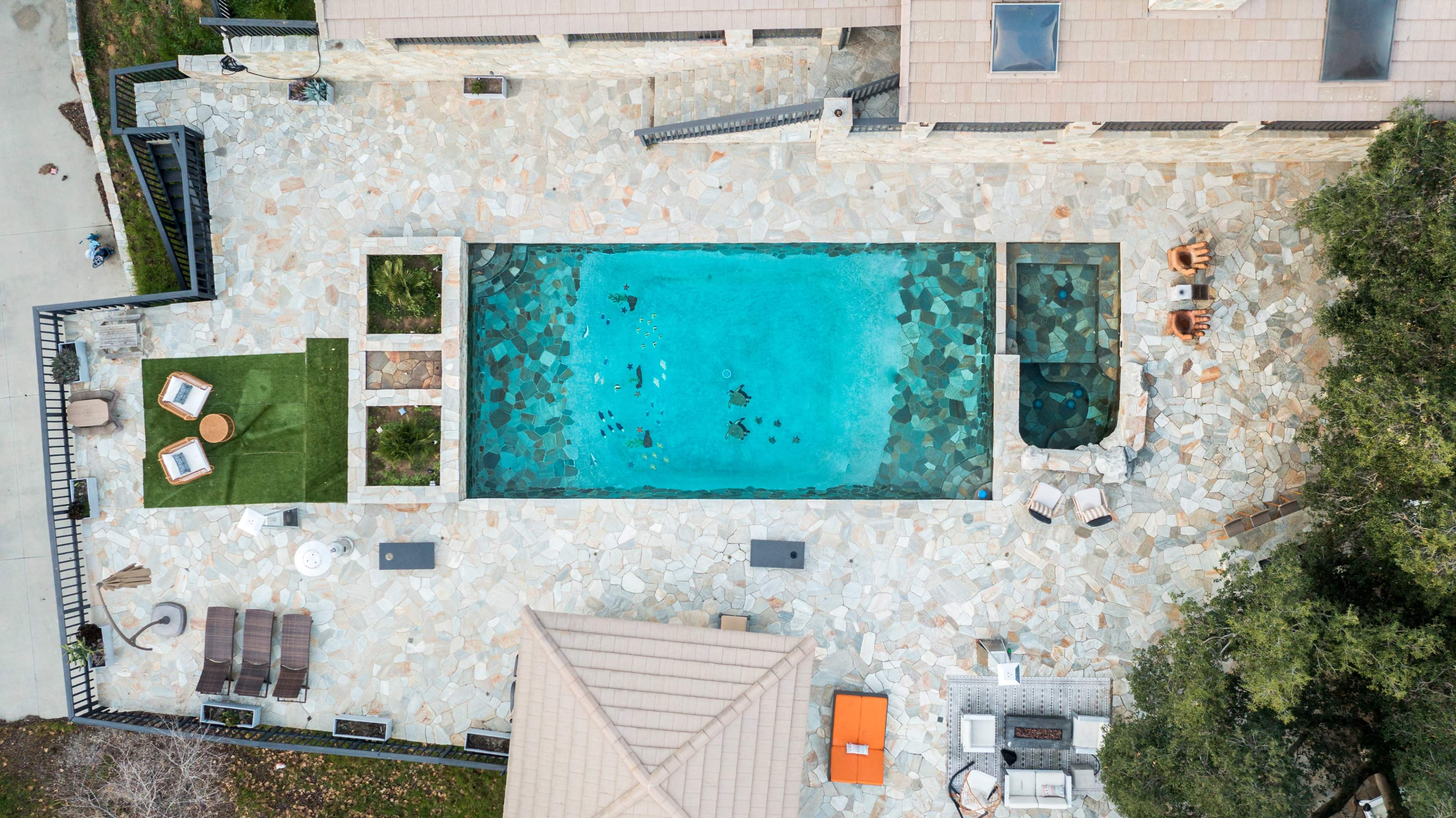 An aerial view shows a stone patio surrounding a rectangular swimming pool with a spa, lounge chairs, and outdoor seating areas.