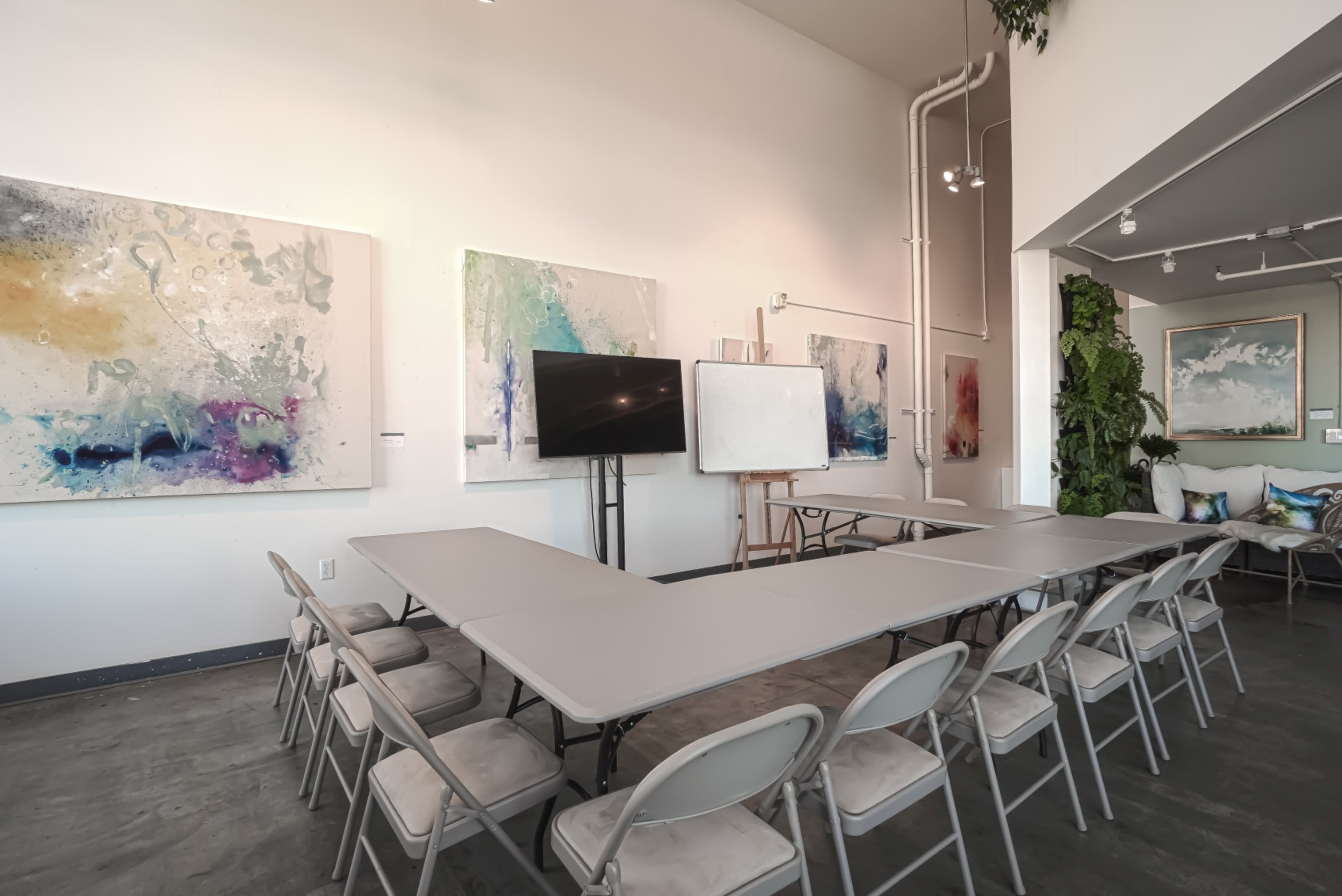 A modern meeting space features a long table surrounded by folding chairs, with large abstract paintings on the walls and a screen for presentations.