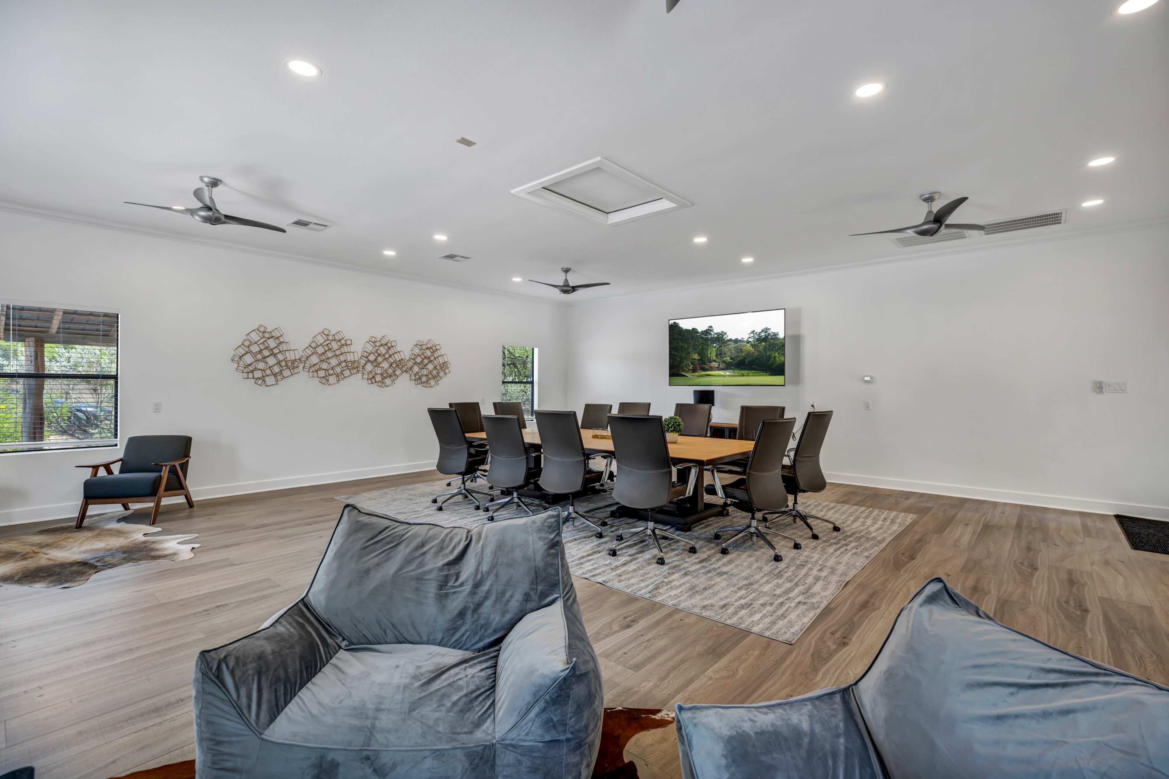A modern conference room features a large table surrounded by ergonomic chairs, with a large screen on one wall and casual seating in the foreground.
