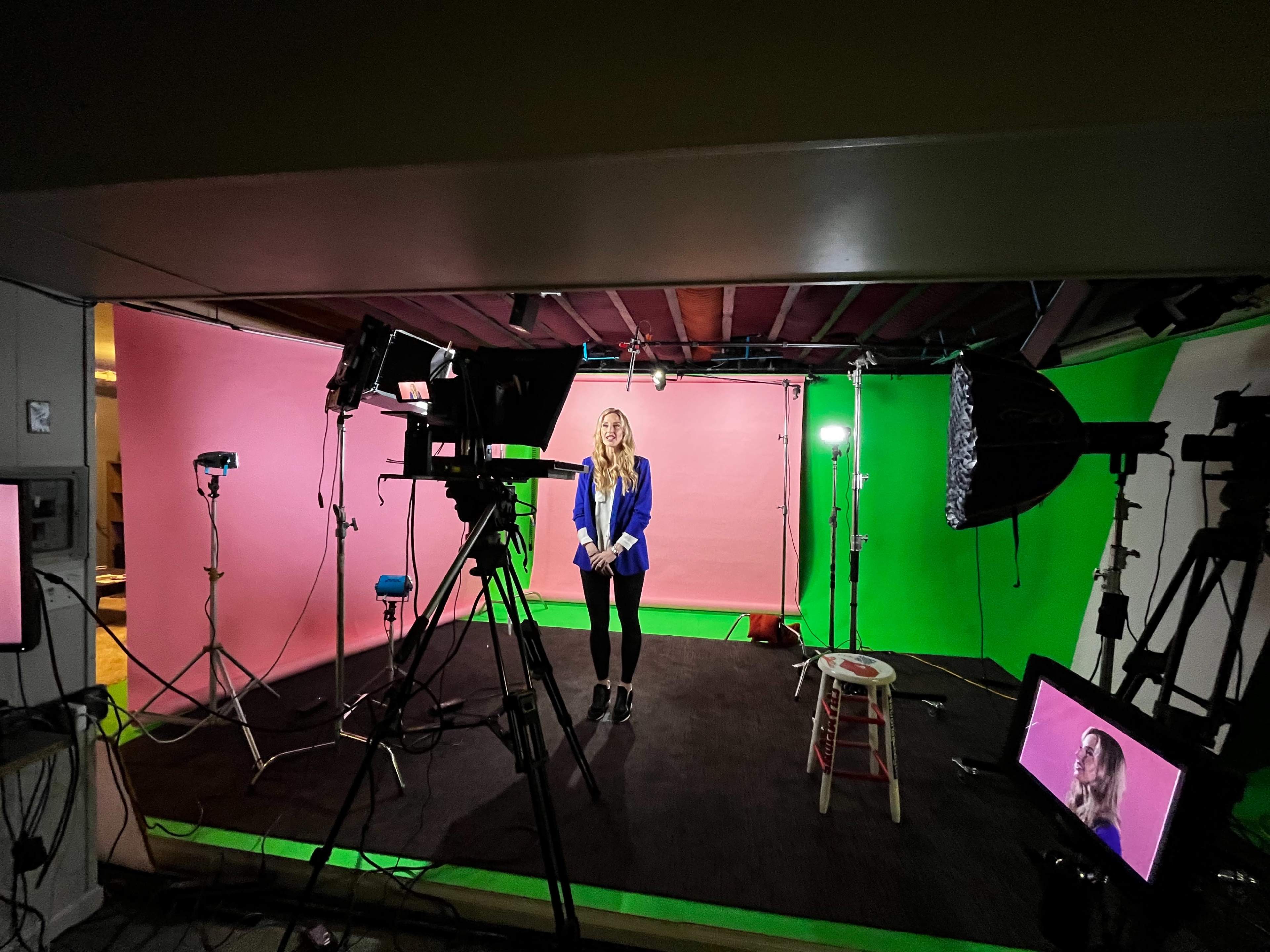 A woman stands in a studio with pink and green backdrop walls, surrounded by multiple cameras and lights.