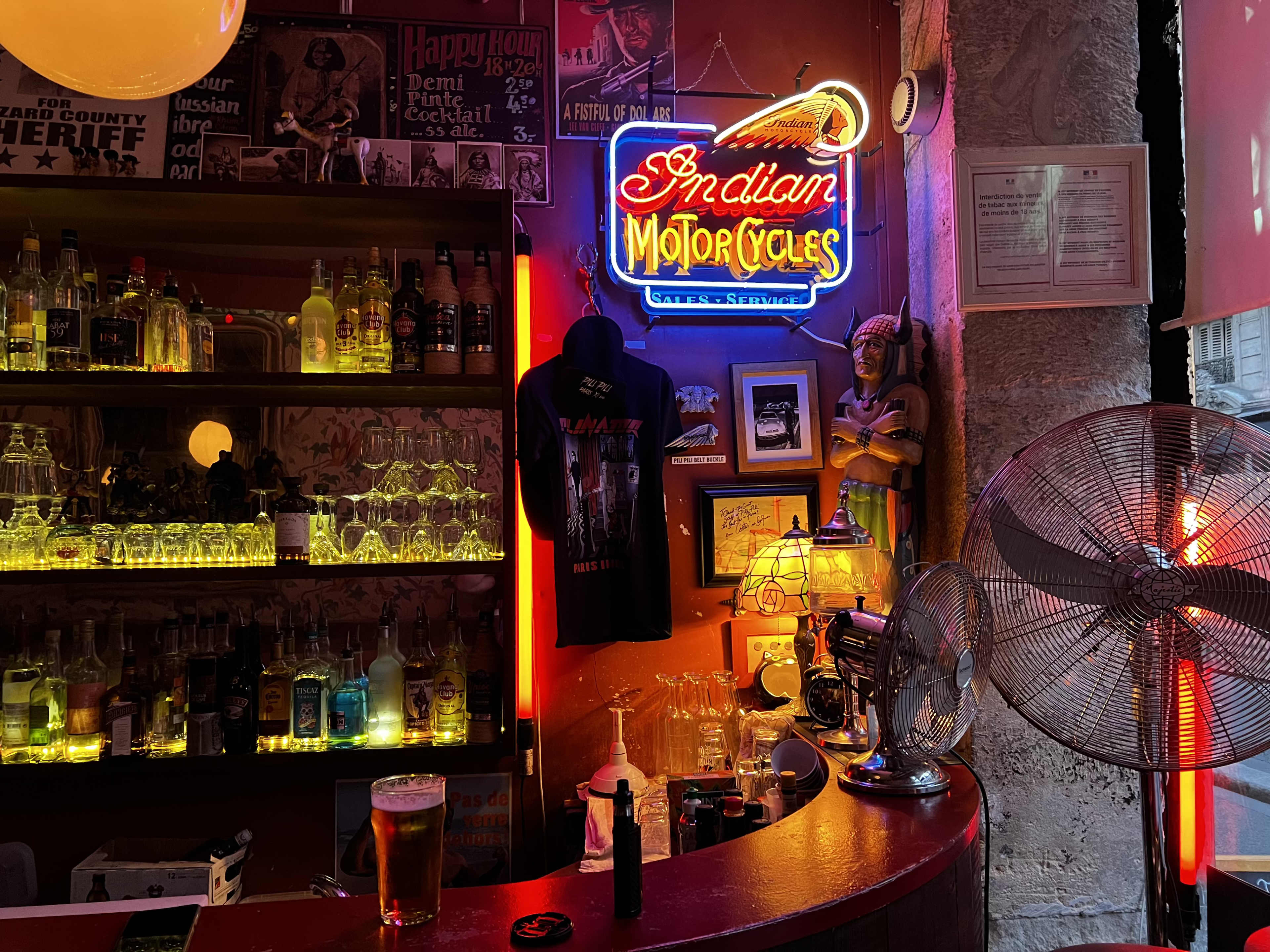 The bar features a neon sign reading "Indian Motorcycles" alongside shelves filled with various bottles and a fan in the background.