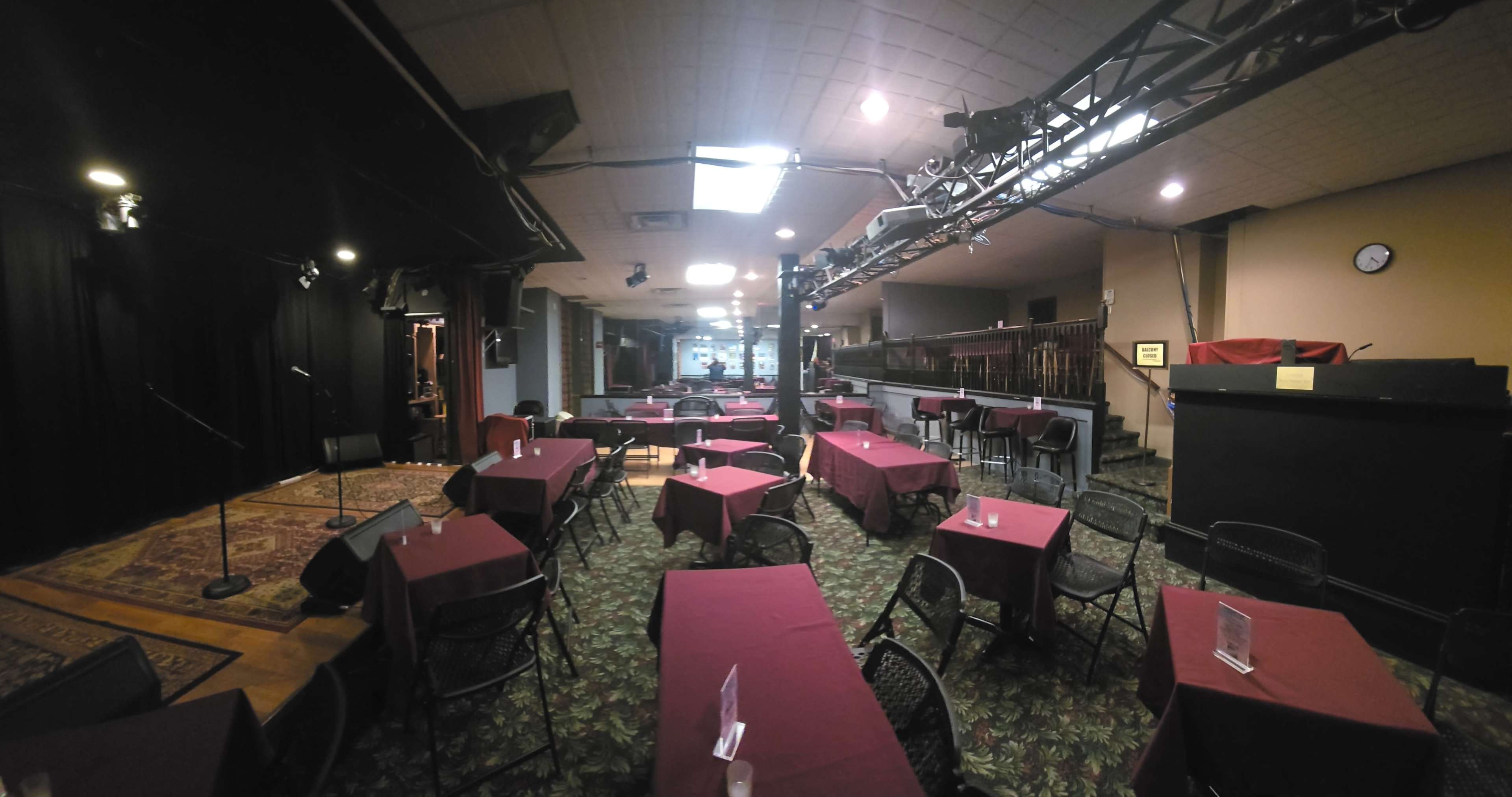 The image shows an empty performance venue with tables covered in red tablecloths and a small stage area in the background.