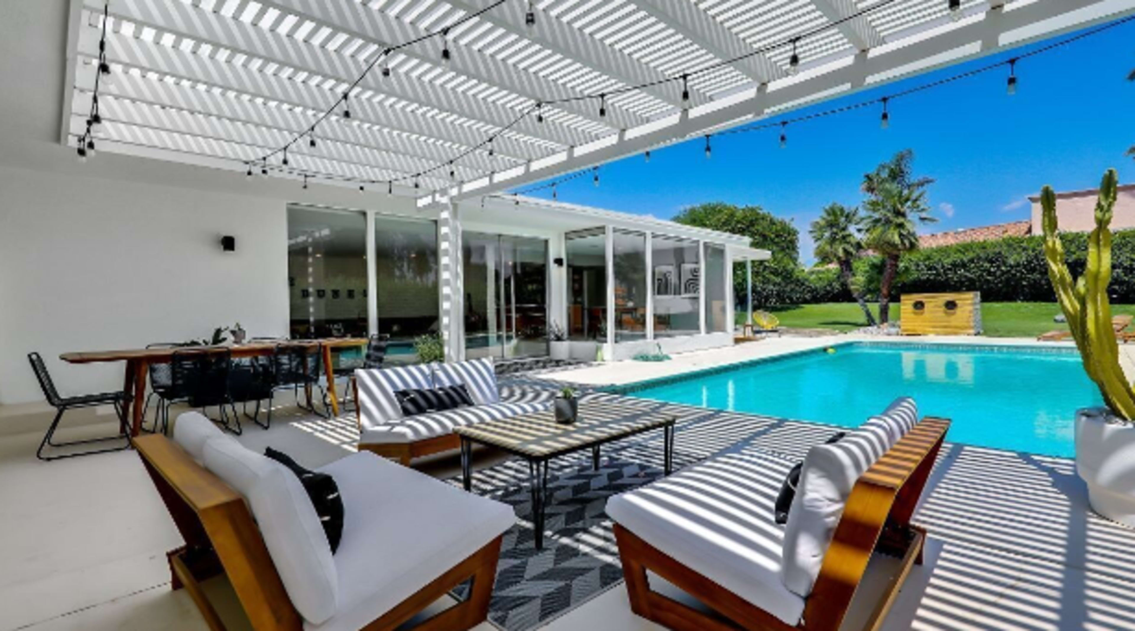 The image shows a modern patio area with lounge chairs and a dining table facing a sparkling pool, surrounded by greenery and a bright sky.