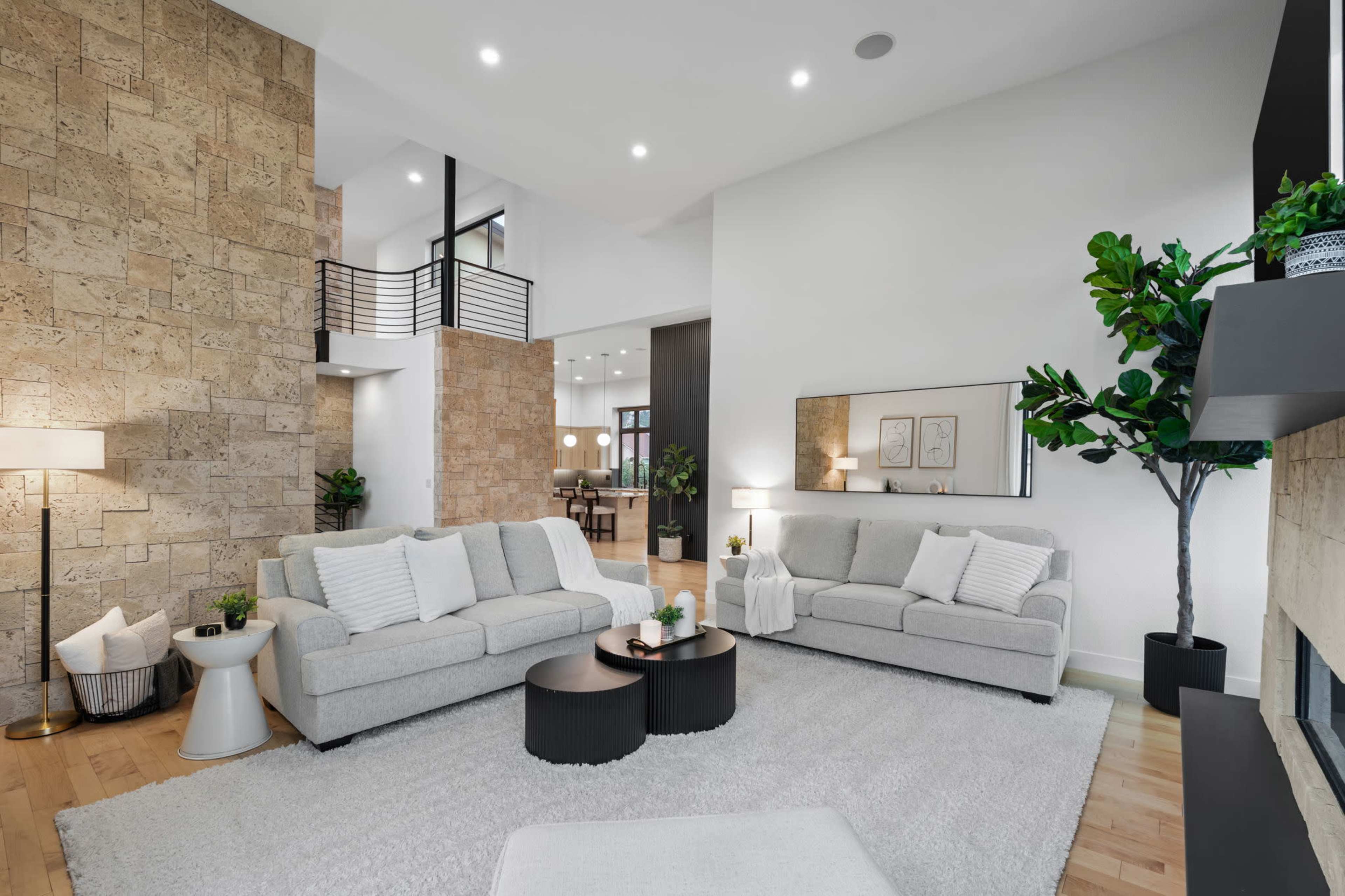The image shows a modern living room with two light gray sofas, a set of black coffee tables, and a beige rug on a wooden floor, surrounded by decorative plants and a stone accent wall.