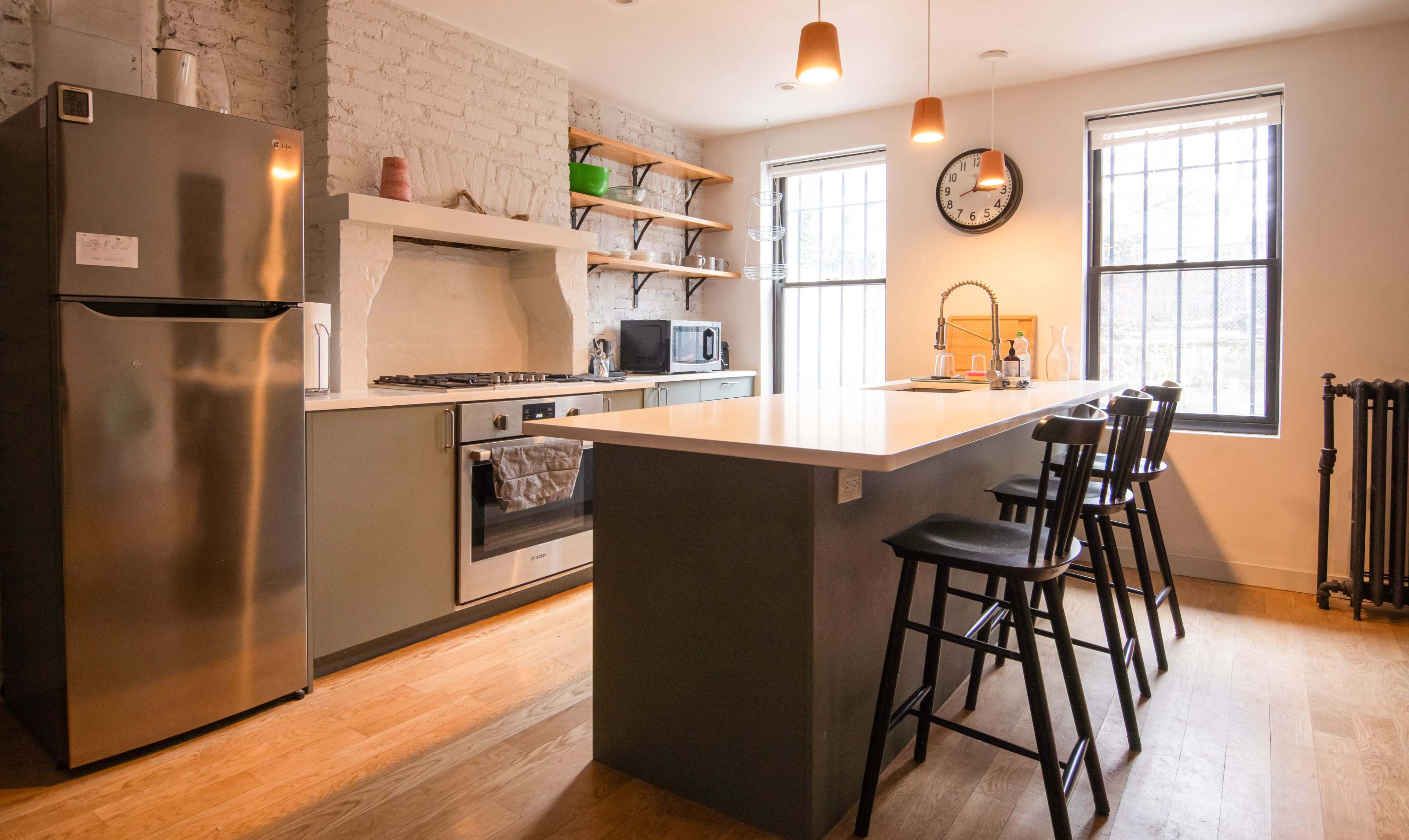 The image shows a modern kitchen featuring a stainless steel refrigerator, stove, and an island with three bar stools, set against a backdrop of exposed brick and wooden shelves.