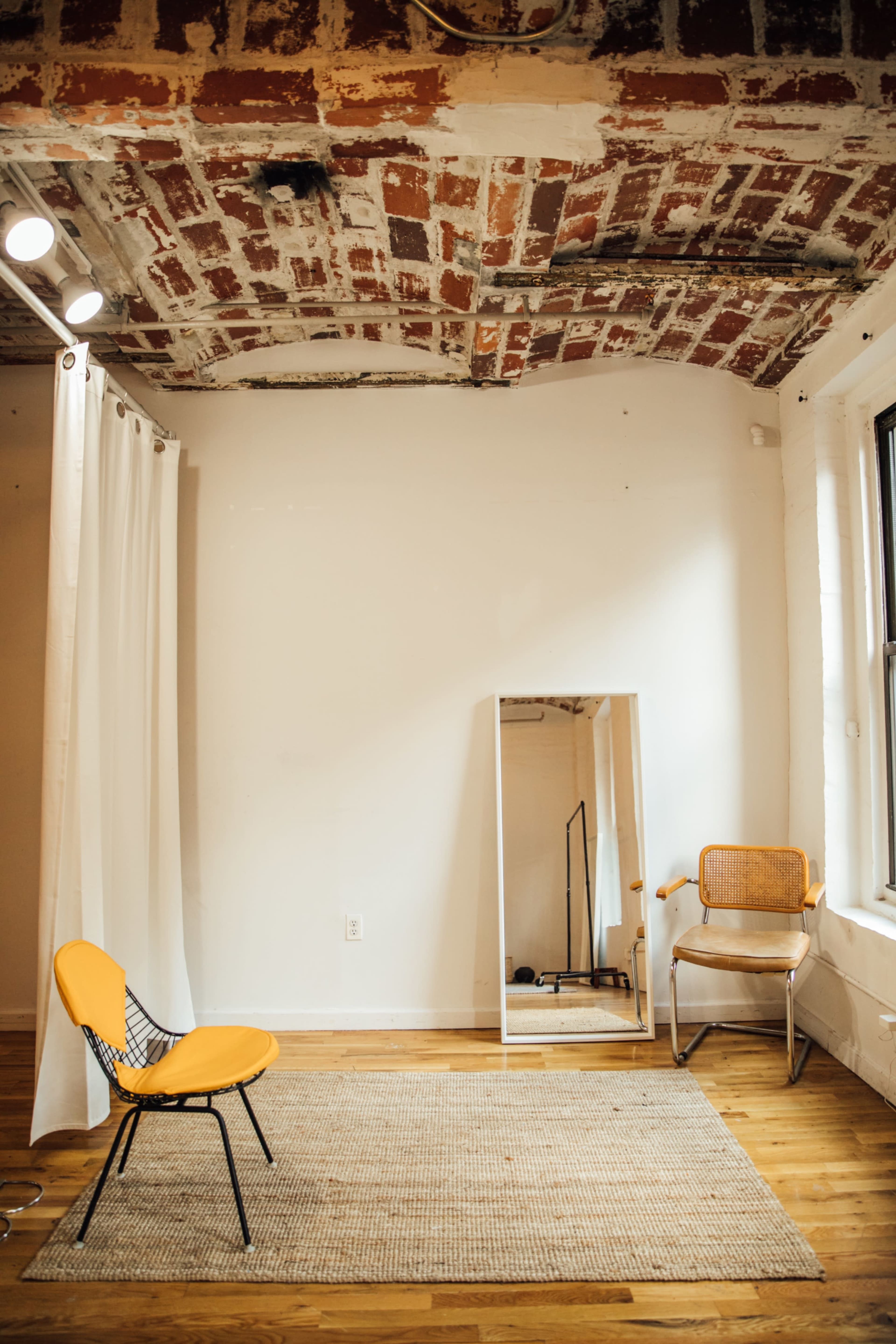 A minimalist room features a yellow chair, a mirror, and a rug on wooden flooring beneath a ceiling with exposed brick.