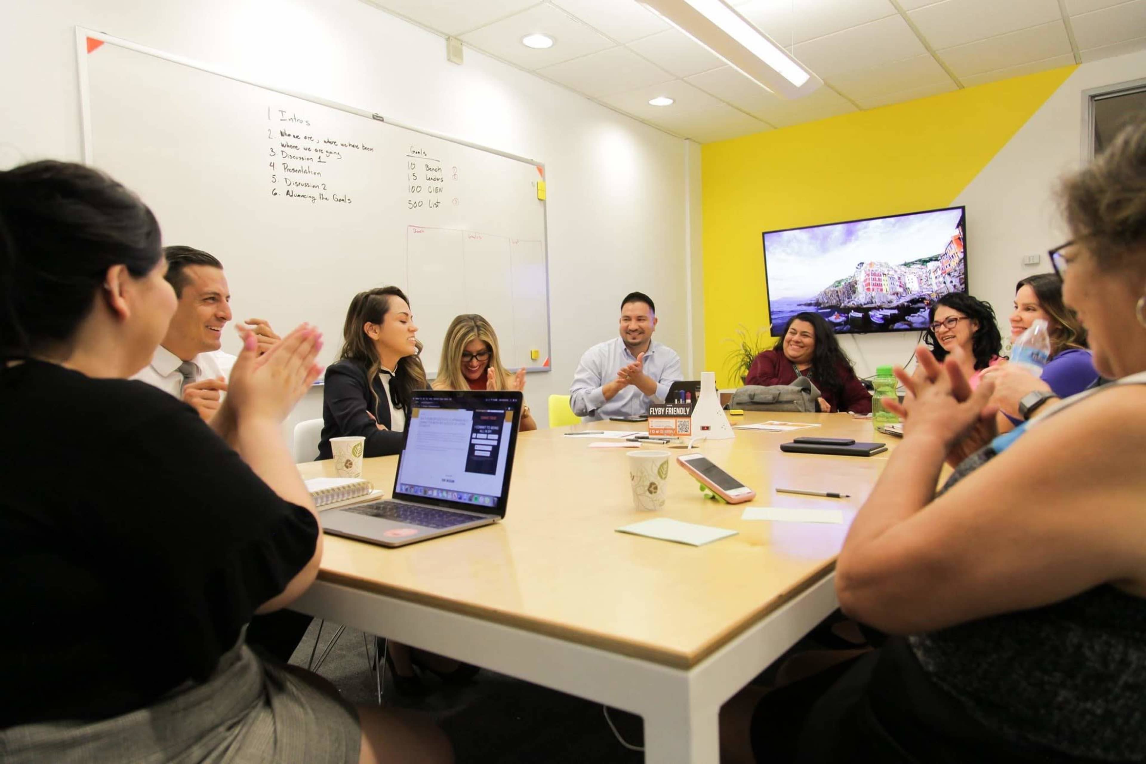 A diverse group of people is seated around a table in a brightly lit conference room, engaging in a collaborative discussion while using laptops and notepads.