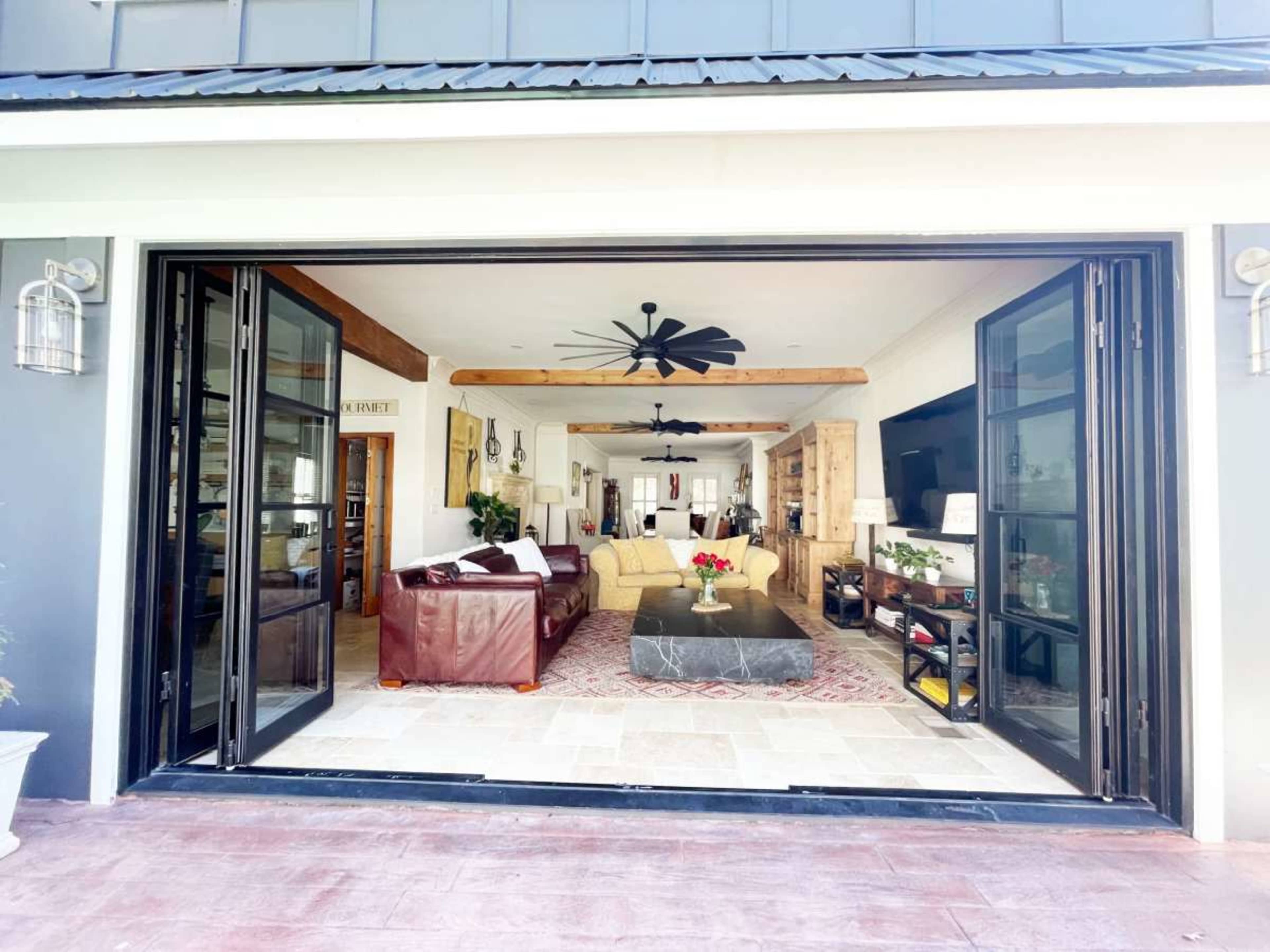 The image shows a spacious living room with large, open doors leading to an outdoor area, featuring a seating arrangement, a coffee table, and decorative elements.