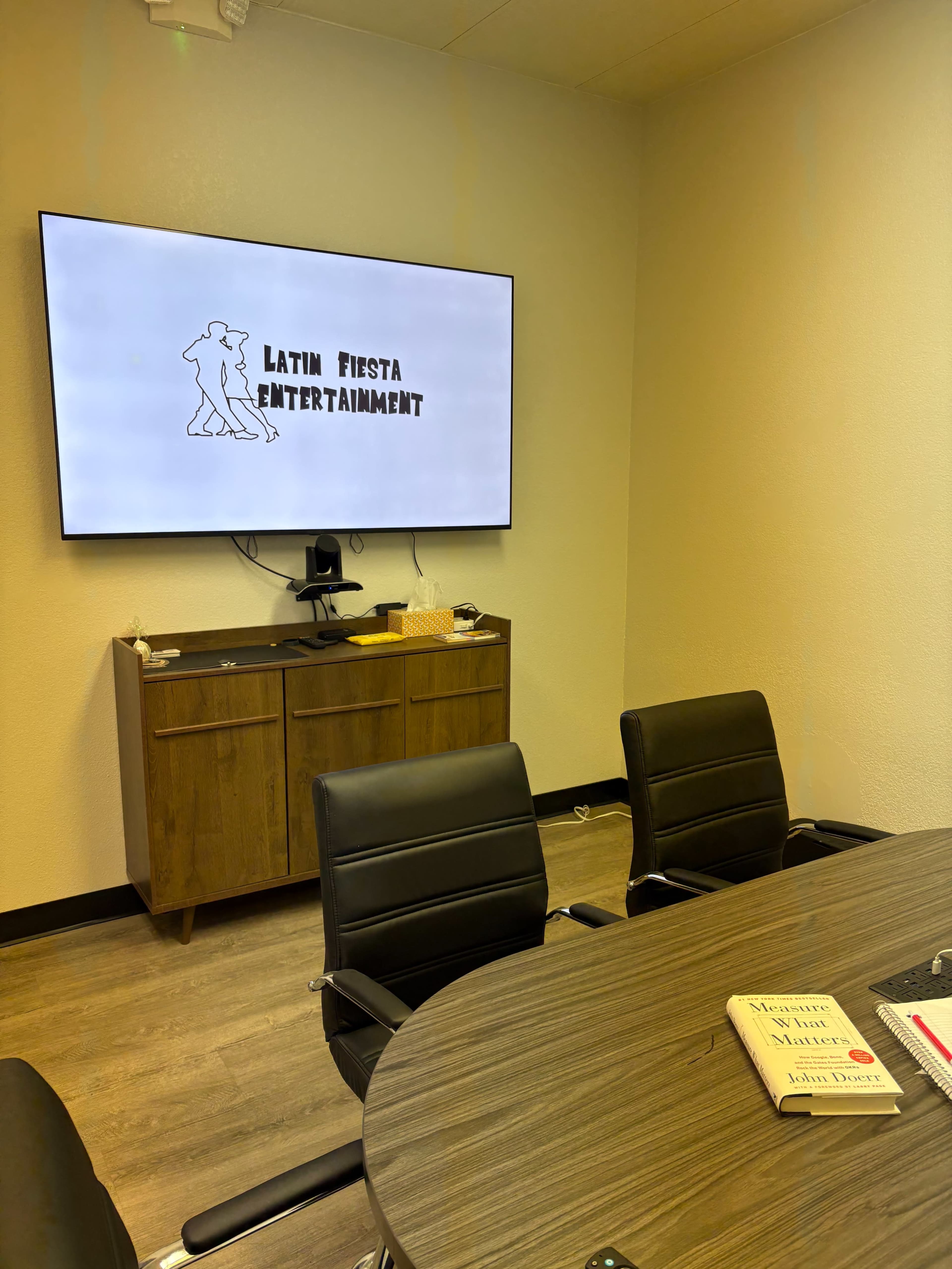 A conference room features a large screen displaying "LATIN FIESTA ENTERTAINMENT" and a wooden table surrounded by black chairs.