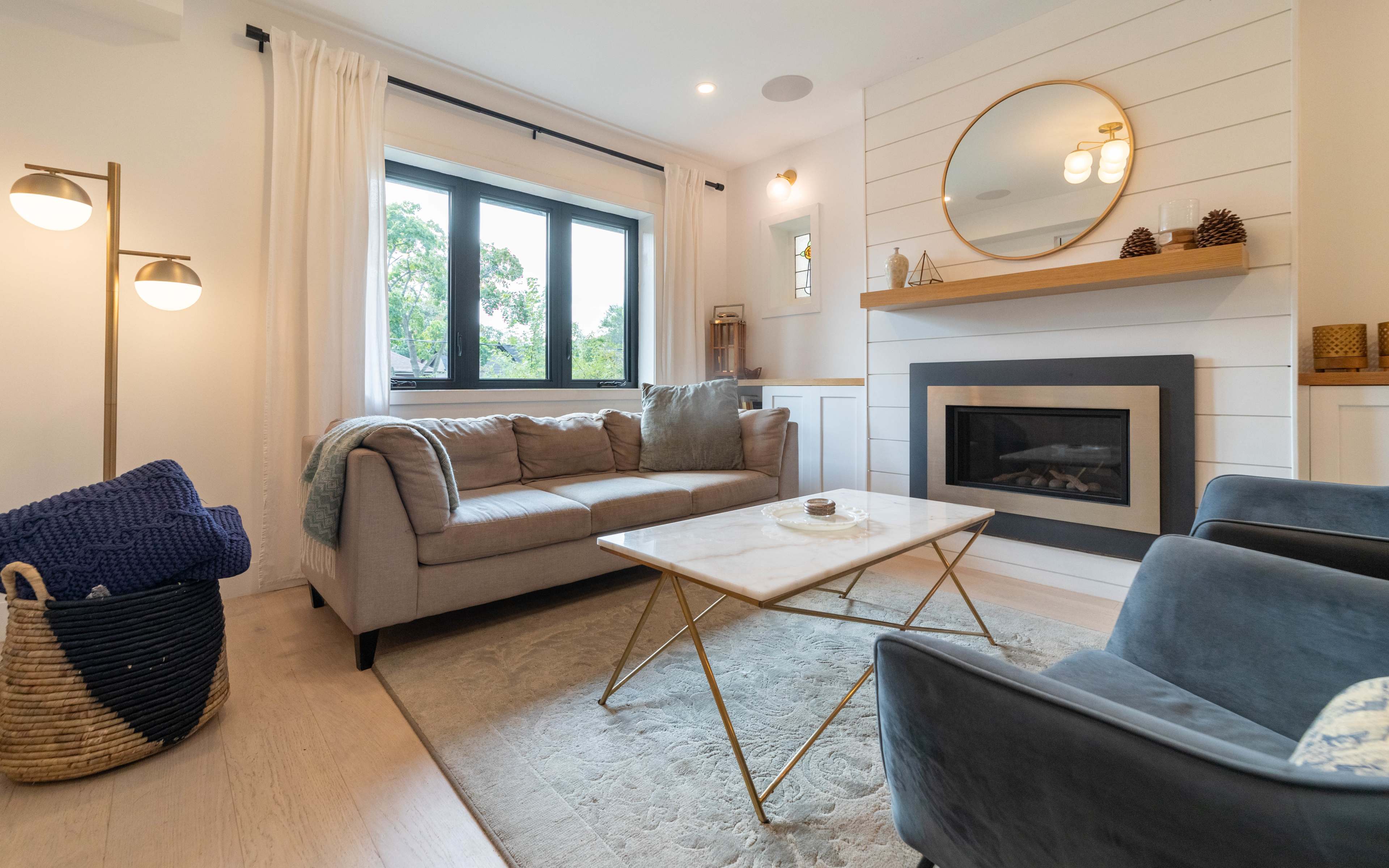 A modern living room with a gray sofa, a marble-top coffee table, a fireplace beneath a wooden mantel, and large windows allowing natural light.