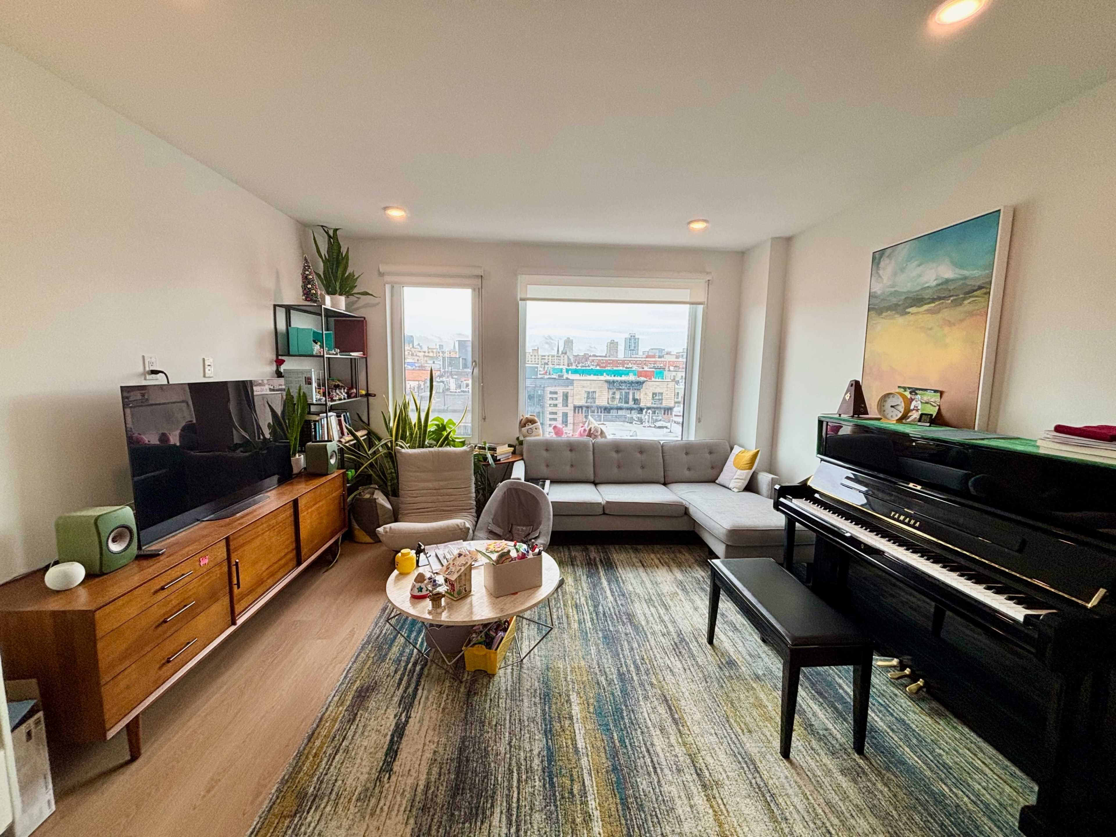 The image shows a modern living room featuring a gray sectional sofa, a black piano, a wooden TV unit, and large windows with city views.