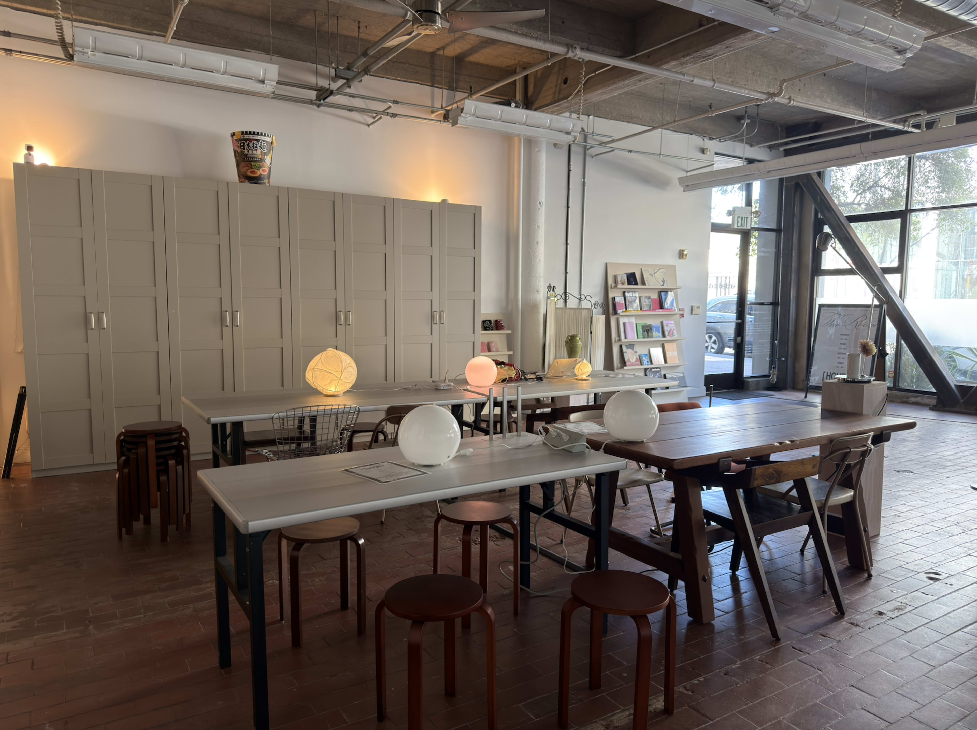 The image shows a spacious, well-lit room with several tables, chairs, and storage cabinets, featuring a mix of decorative lamps and shelves containing books.