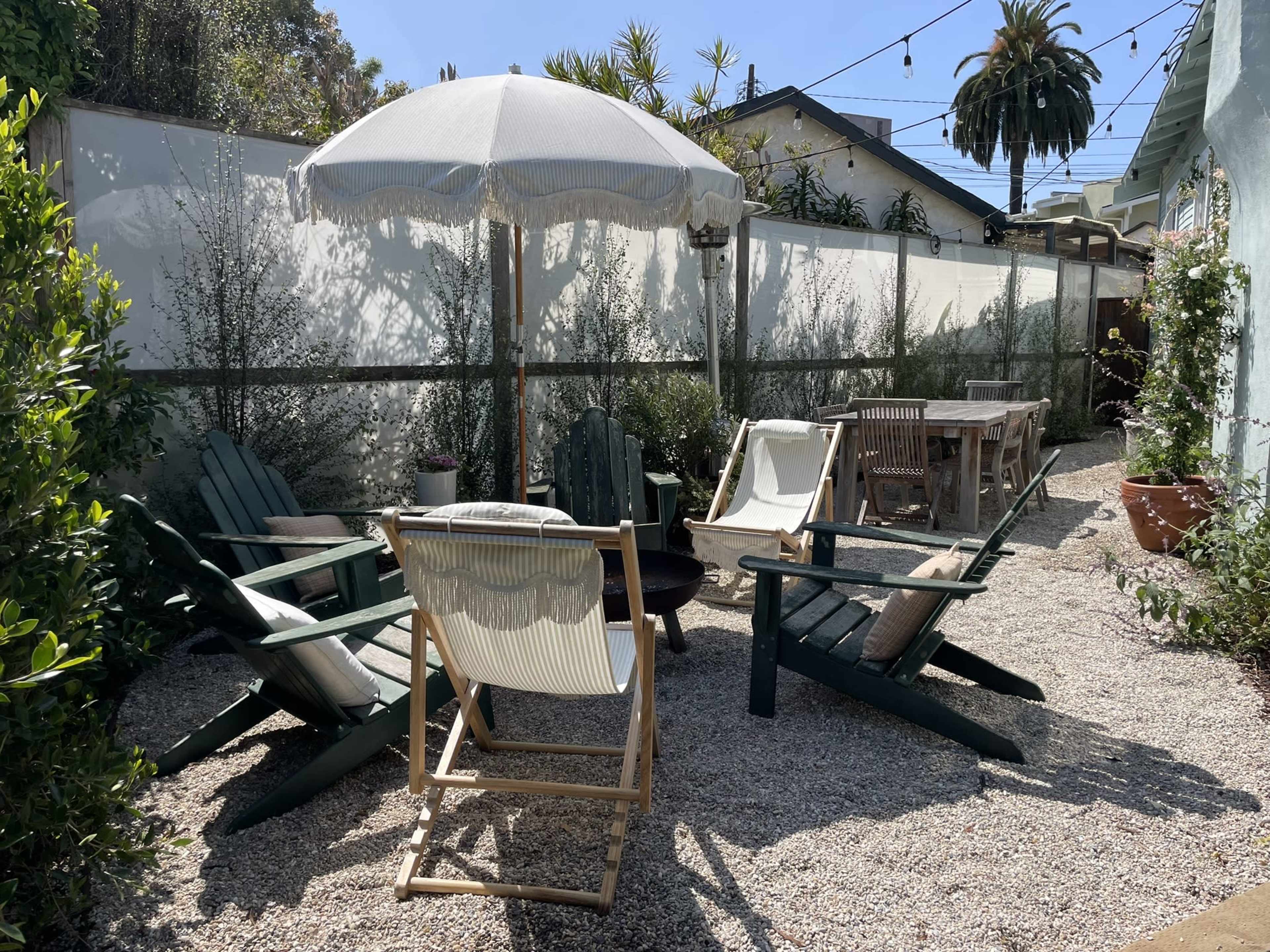 The scene shows a gravel outdoor area with several chairs arranged around a fire pit, a large umbrella for shade, and a dining table in the background, surrounded by greenery and a wall.