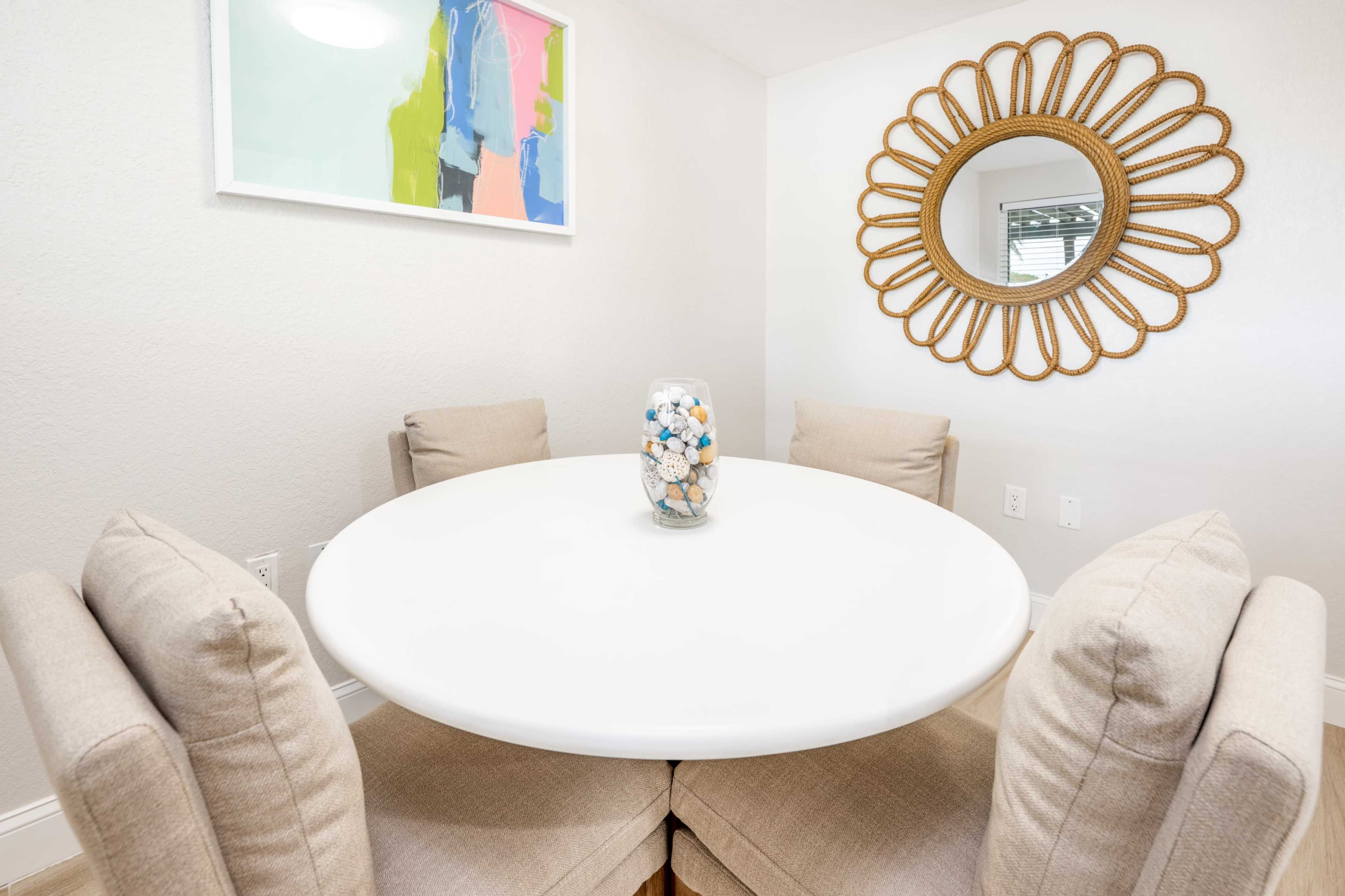A round dining table is set against a light-colored wall, surrounded by upholstered chairs, with a decorative mirror and a colorful artwork above it.