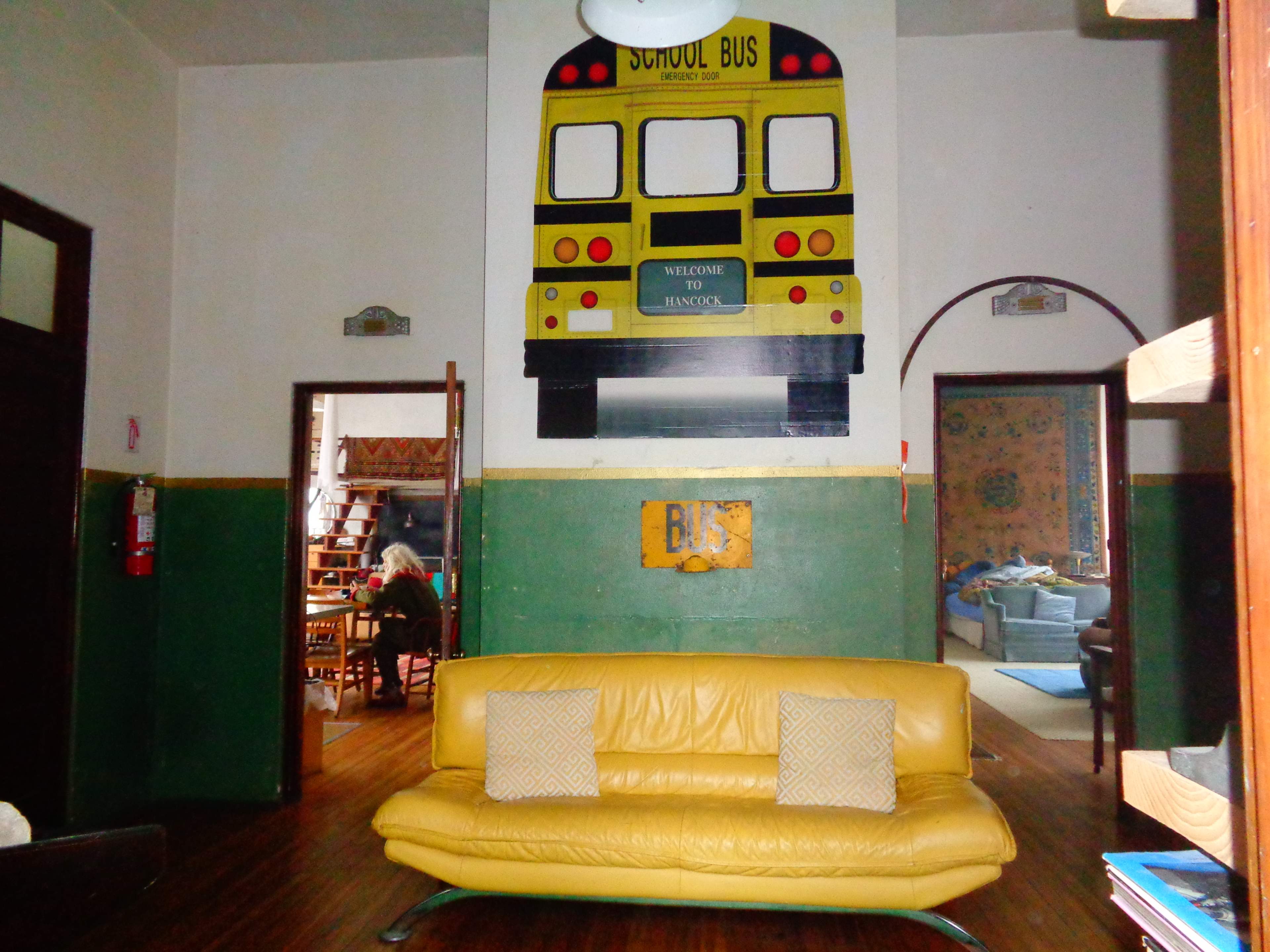 The interior of a room features a large mural of a yellow school bus on the wall above a yellow sofa with two decorative pillows.