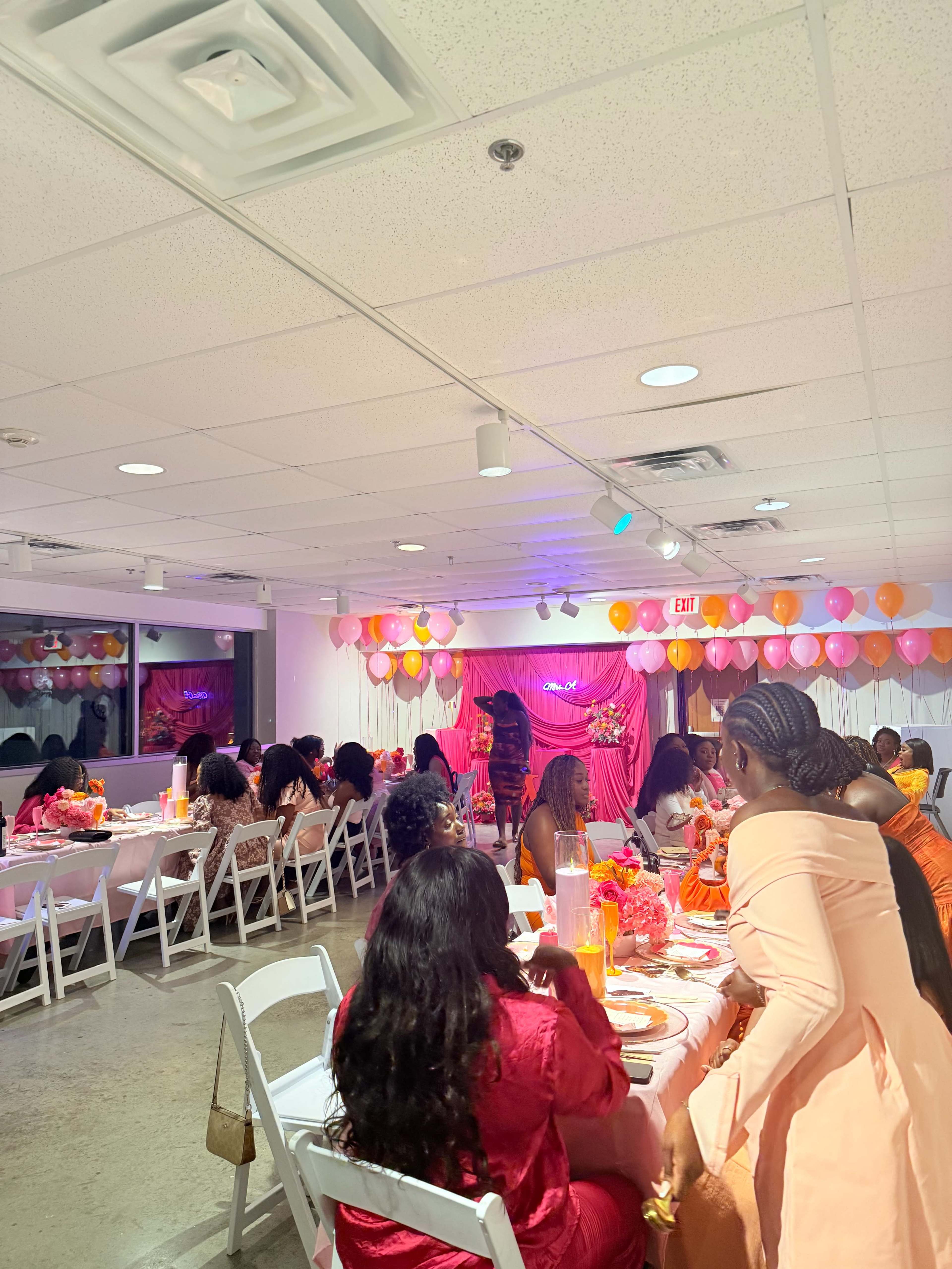 The image shows a well-decorated indoor event space set up for a gathering, with tables arranged for dining and colorful balloons adorning the walls.