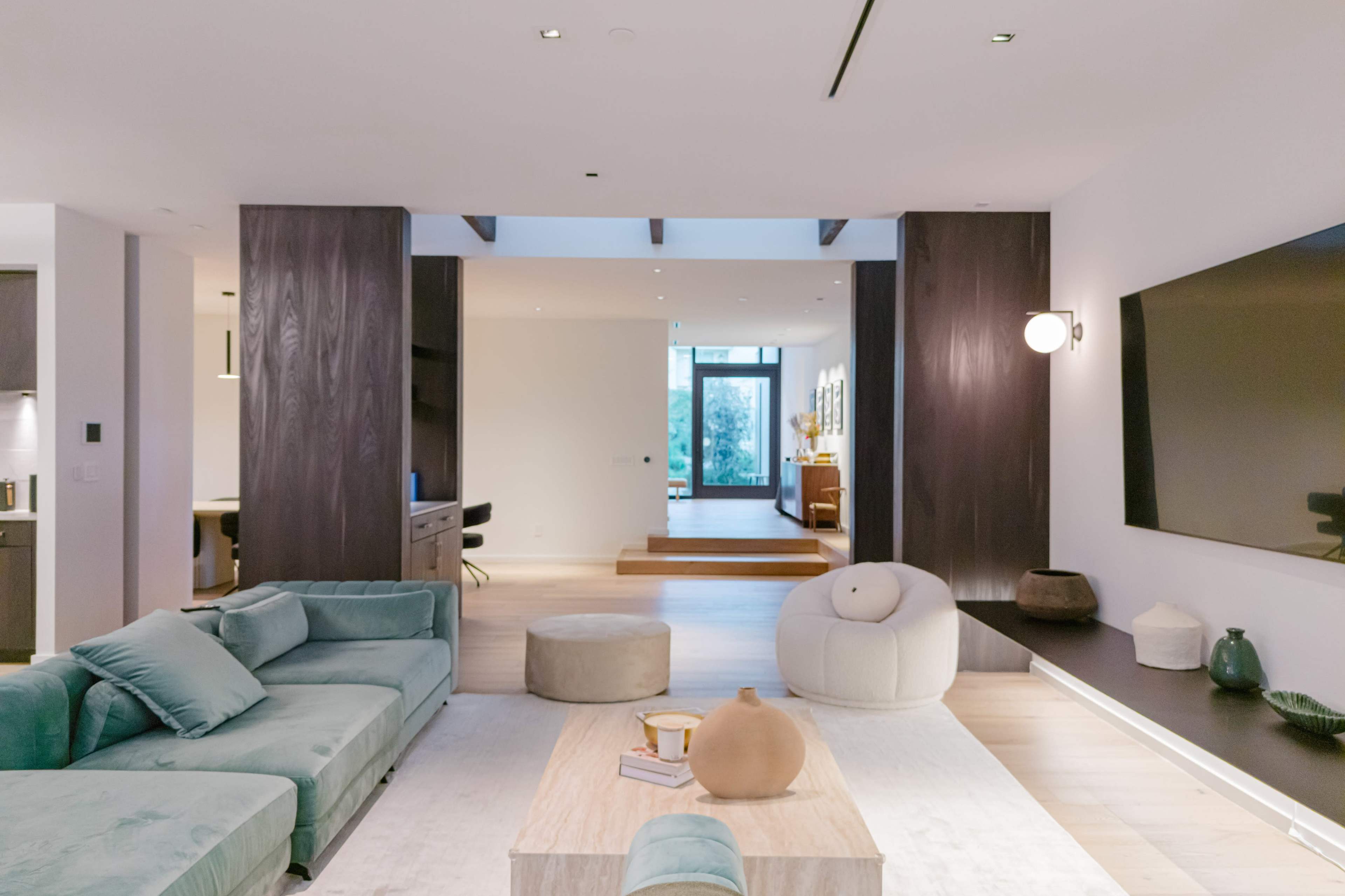 A modern living room features a large green sectional sofa, a wooden coffee table, and a minimalist design with natural light streaming through an open doorway.