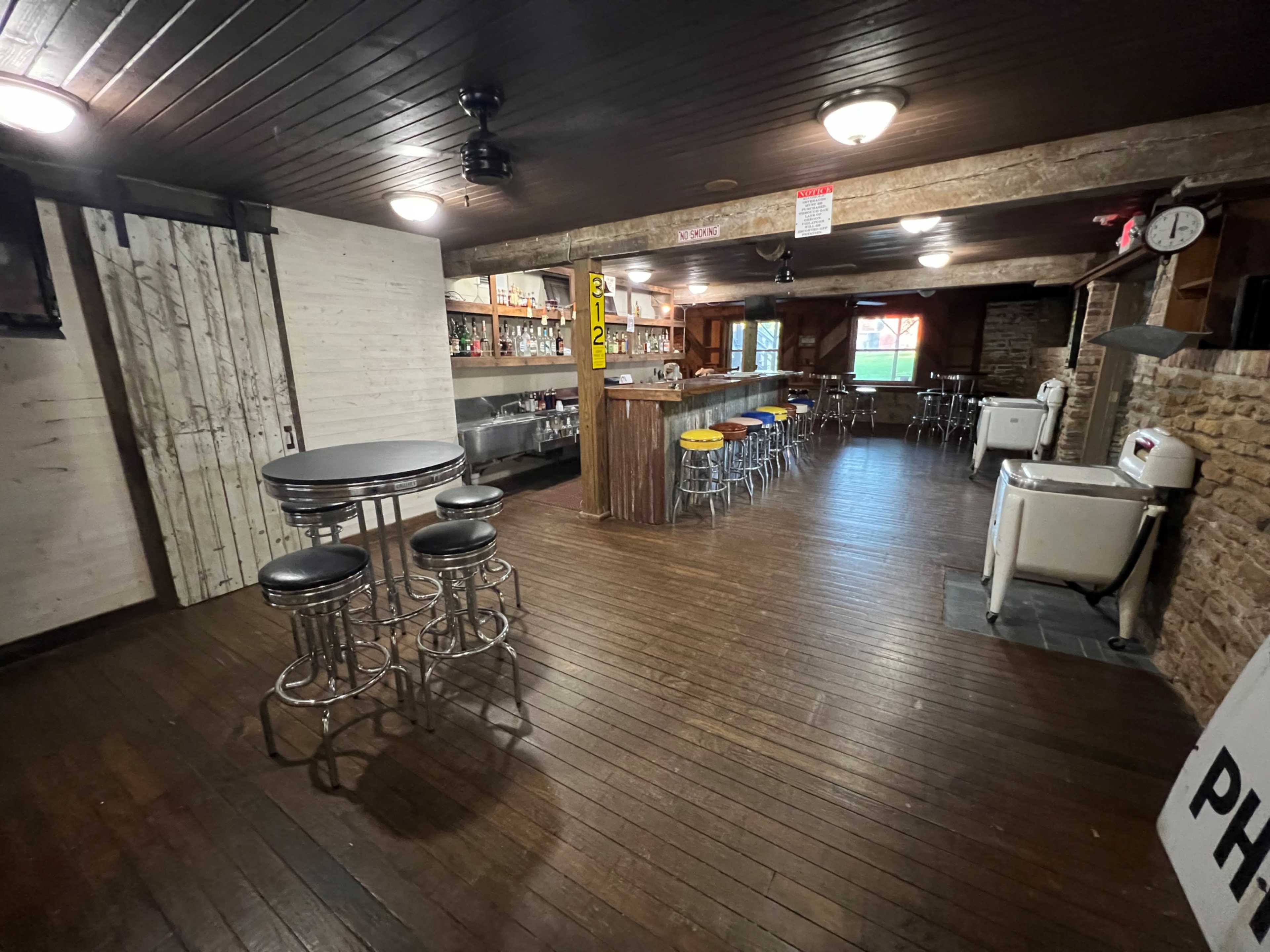 The image shows a rustic, dimly lit bar interior with wooden floors, metal stools, and a long counter lined with shelves of various drink bottles.