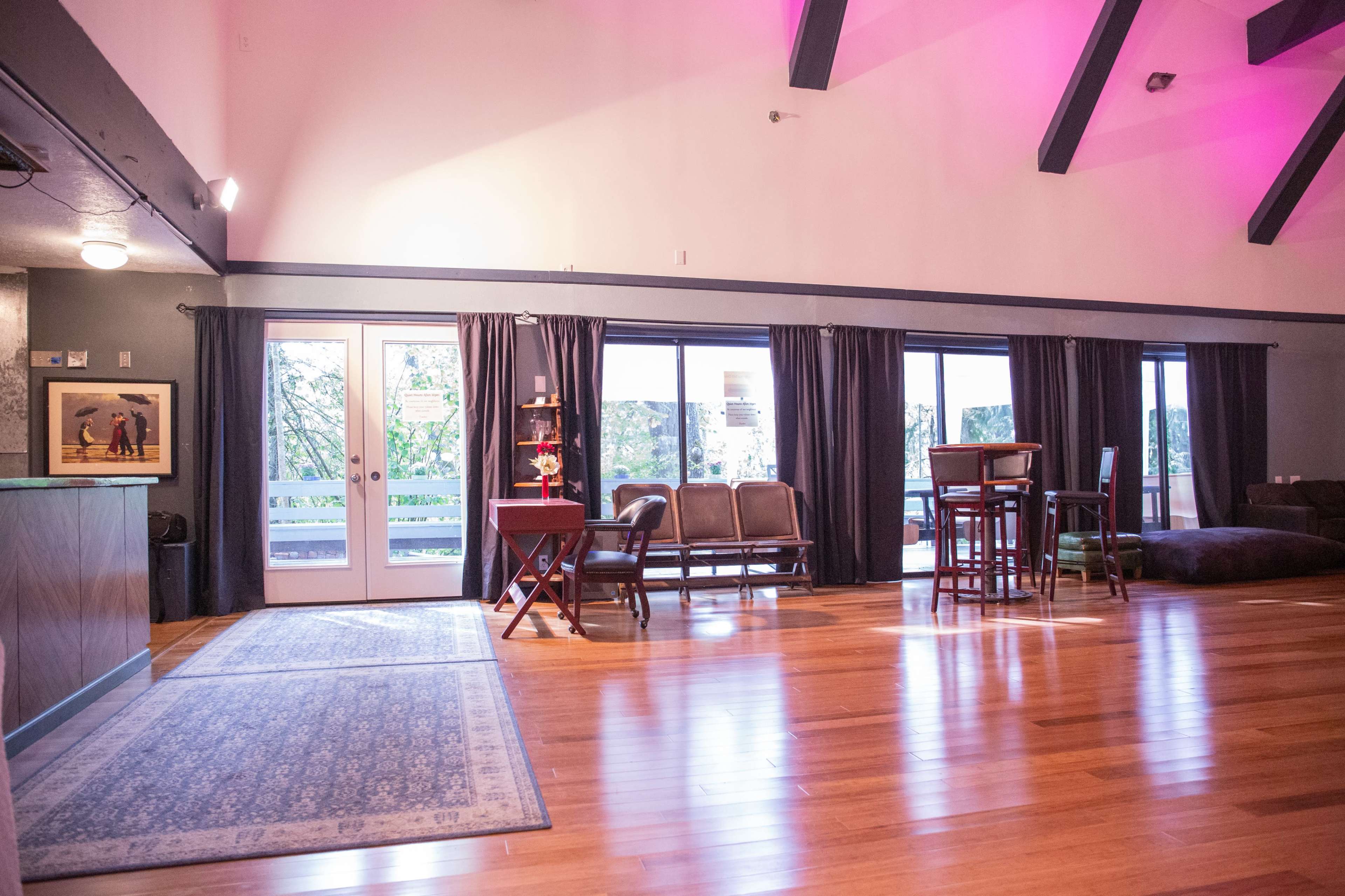 A spacious room with wooden flooring, large windows, a seating area, and a dining table under a vaulted ceiling with beams.