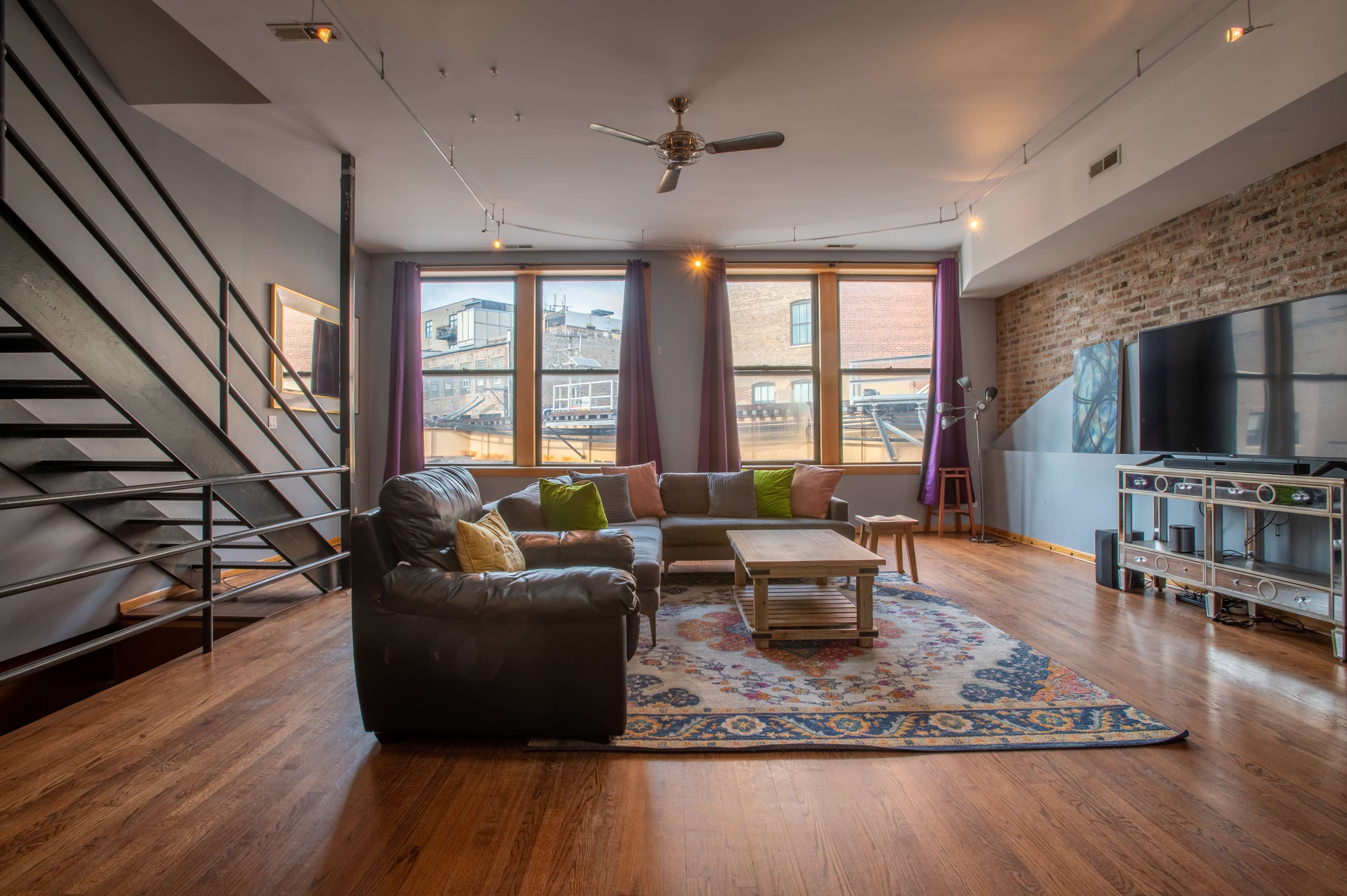 The image shows a spacious living room featuring a black leather sectional sofa, a wooden coffee table on a patterned rug, and large windows with views of a cityscape.