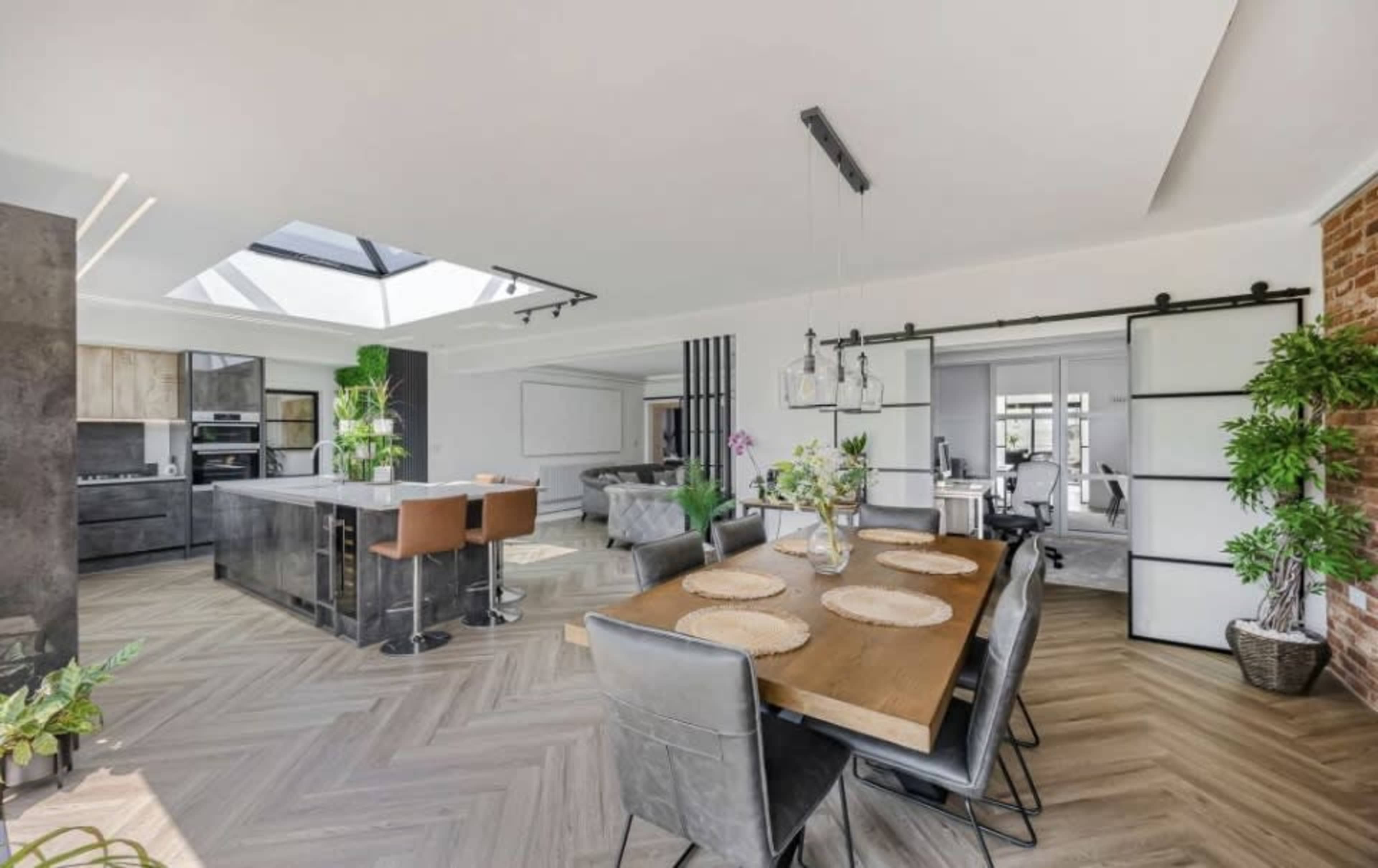 The image shows a modern open-plan kitchen and dining area with a large island, a dining table, and natural light coming through a skylight.