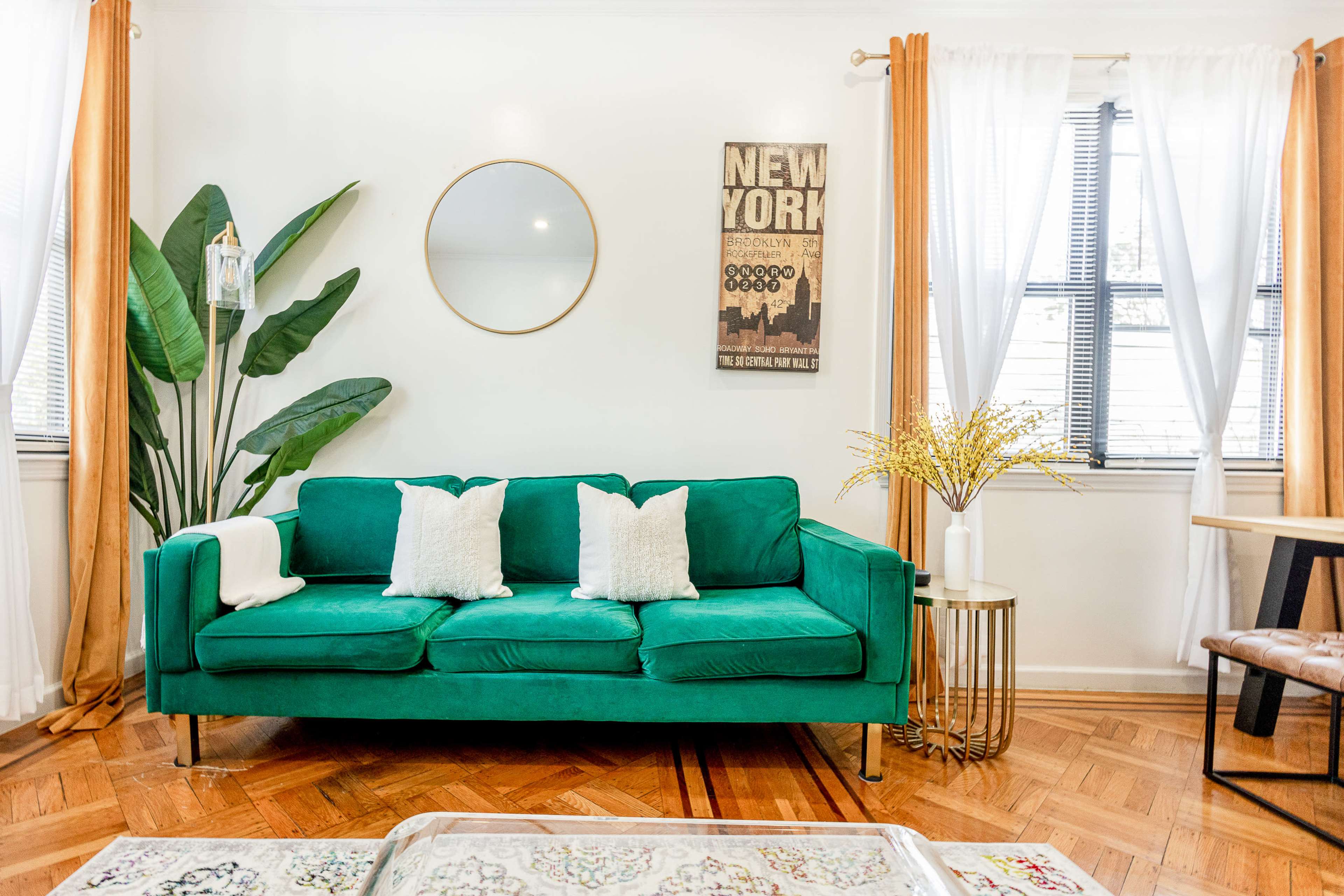 A green sofa with white pillows is positioned in a brightly lit room featuring plants, a round mirror, and decorative curtains.