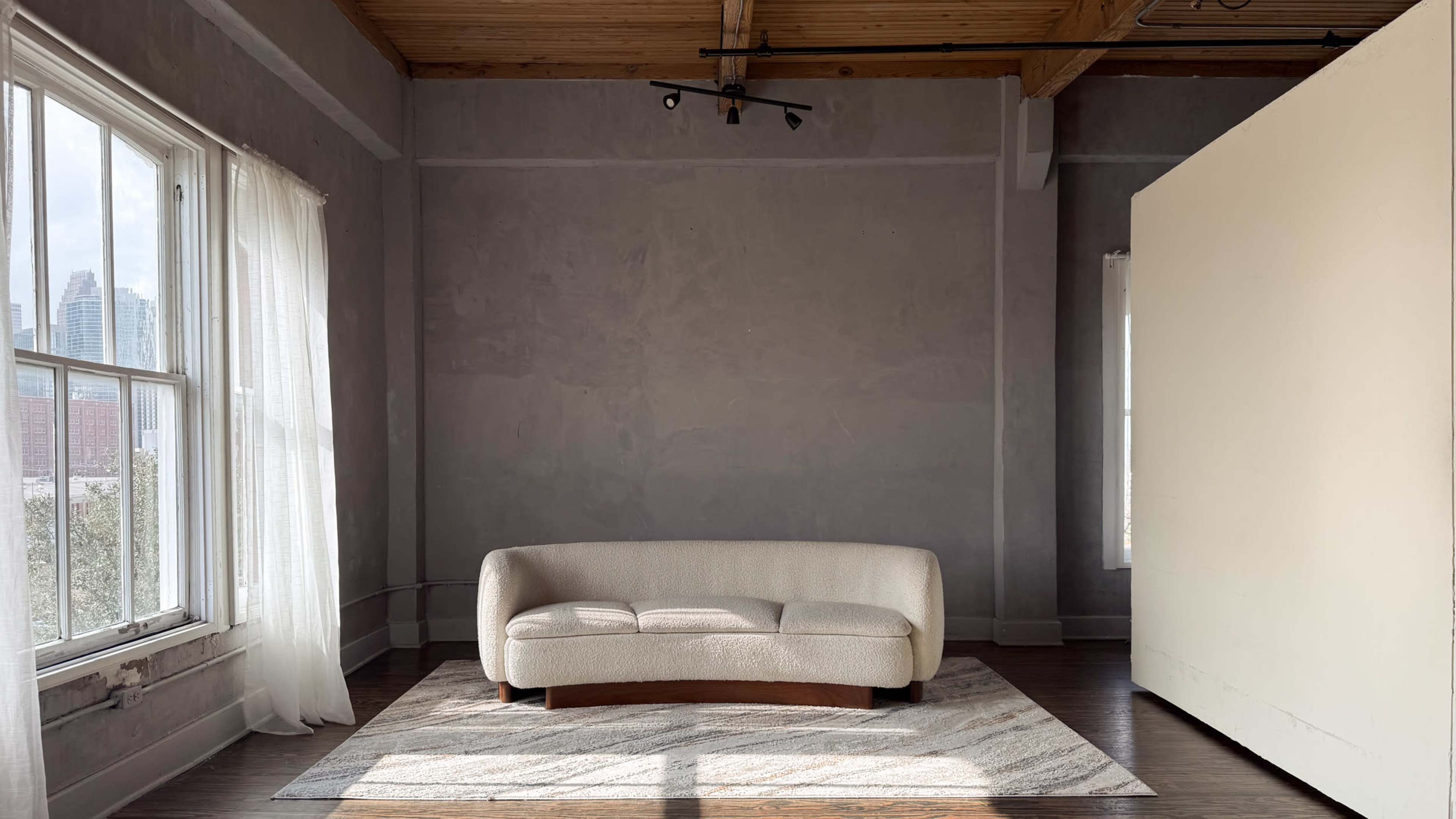 A neutral-toned room features a light-colored sofa on a striped rug, with large windows allowing natural light to fill the space.