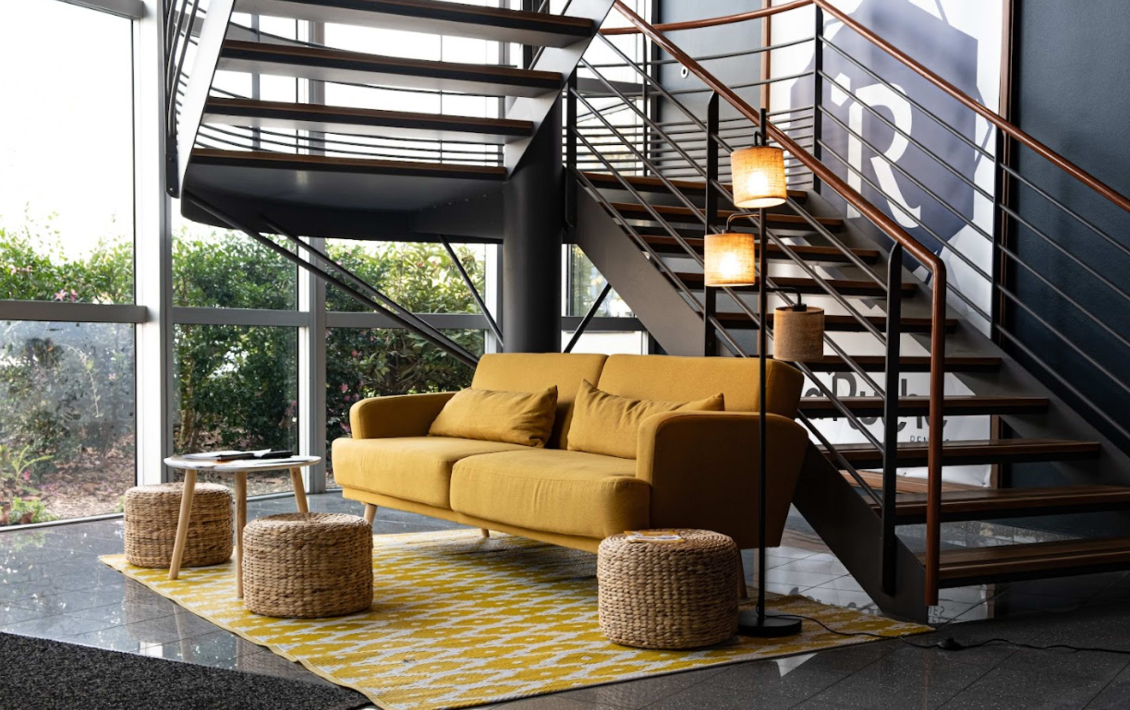 A yellow sofa is placed next to a table and two woven stools on a patterned rug in a space with large windows and a spiral staircase.
