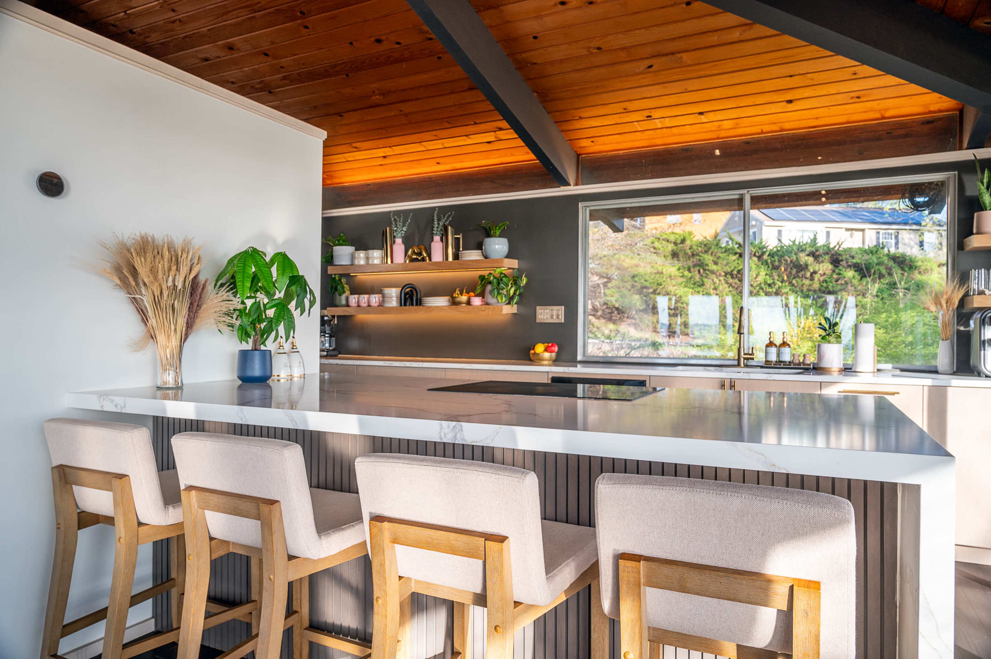 A modern kitchen with a large island bar, four stools, and a large window overlooking a green landscape.