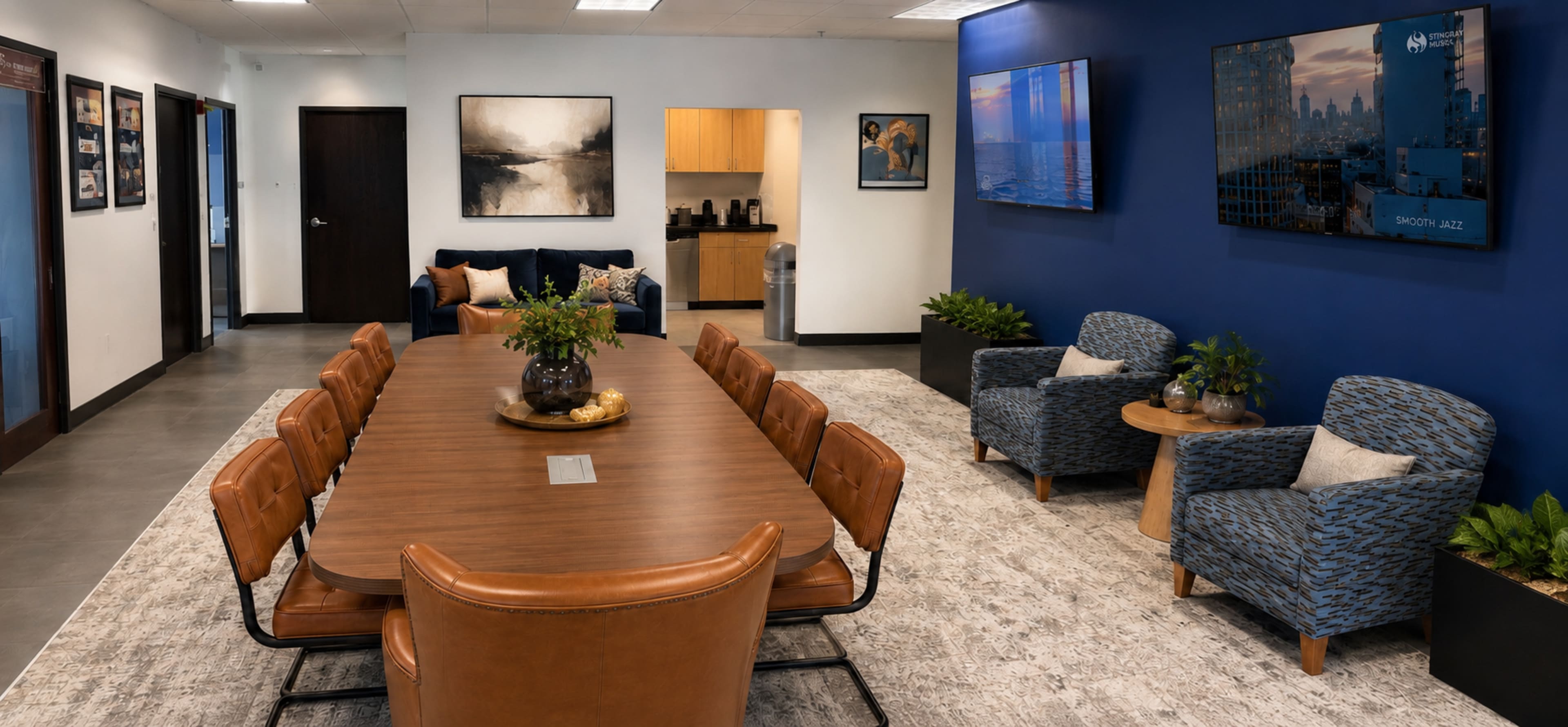 The image shows a modern conference room with a large wooden table, leather chairs, and blue accent walls, featuring a seating area with patterned chairs and art on the walls.