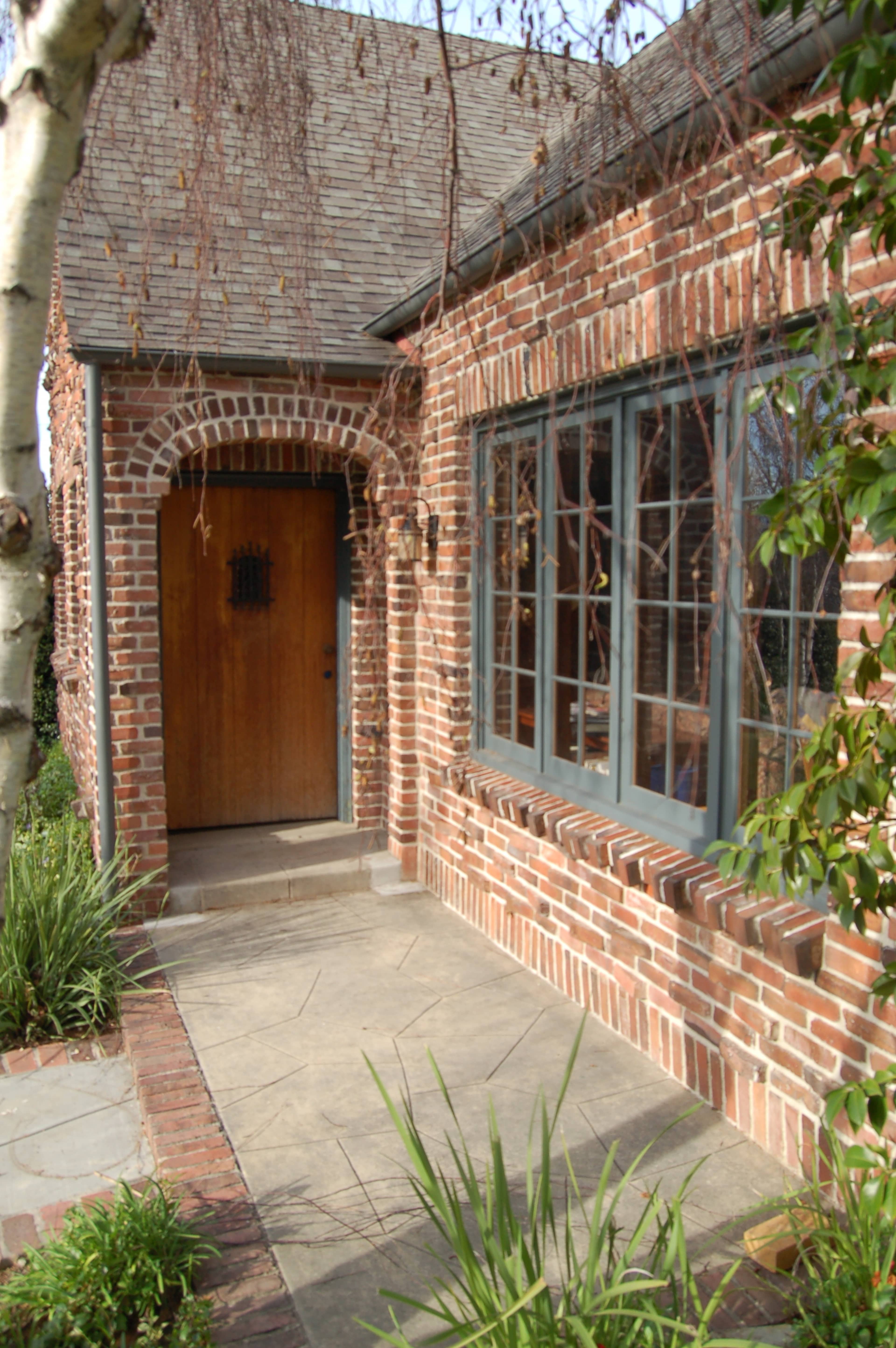 A brick house features a wooden front door and large windows, with a concrete pathway and greenery along the side.