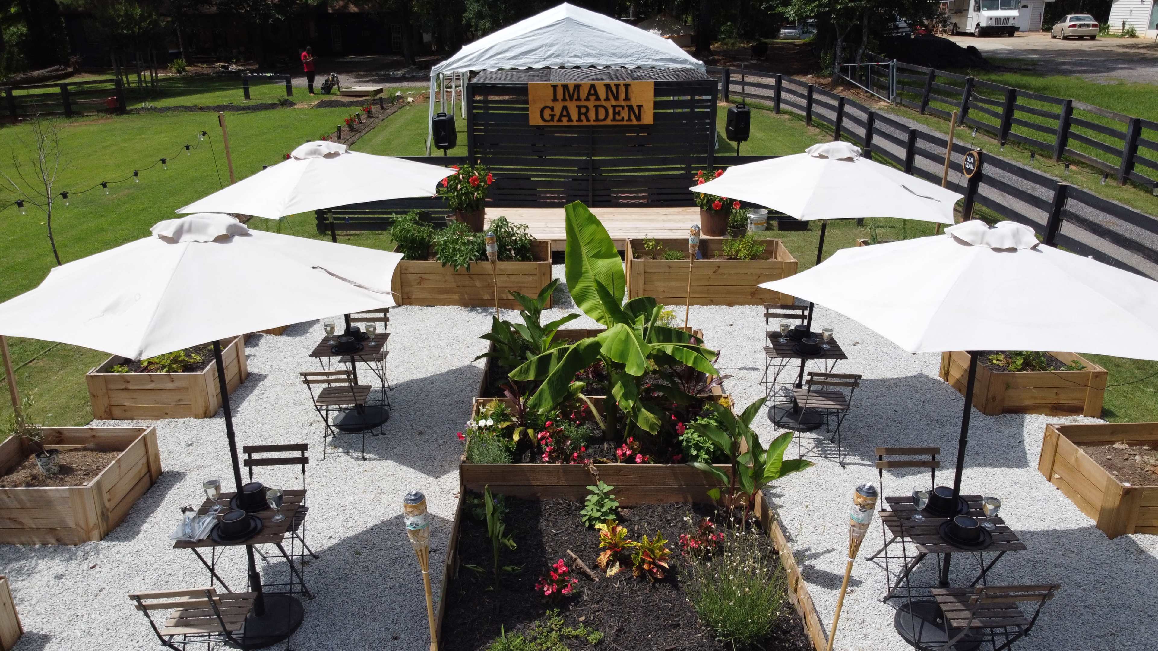 A garden space called "IMANI GARDEN," featuring wooden planters, tables with chairs under white umbrellas, and decorative plants arranged in a gravel layout.