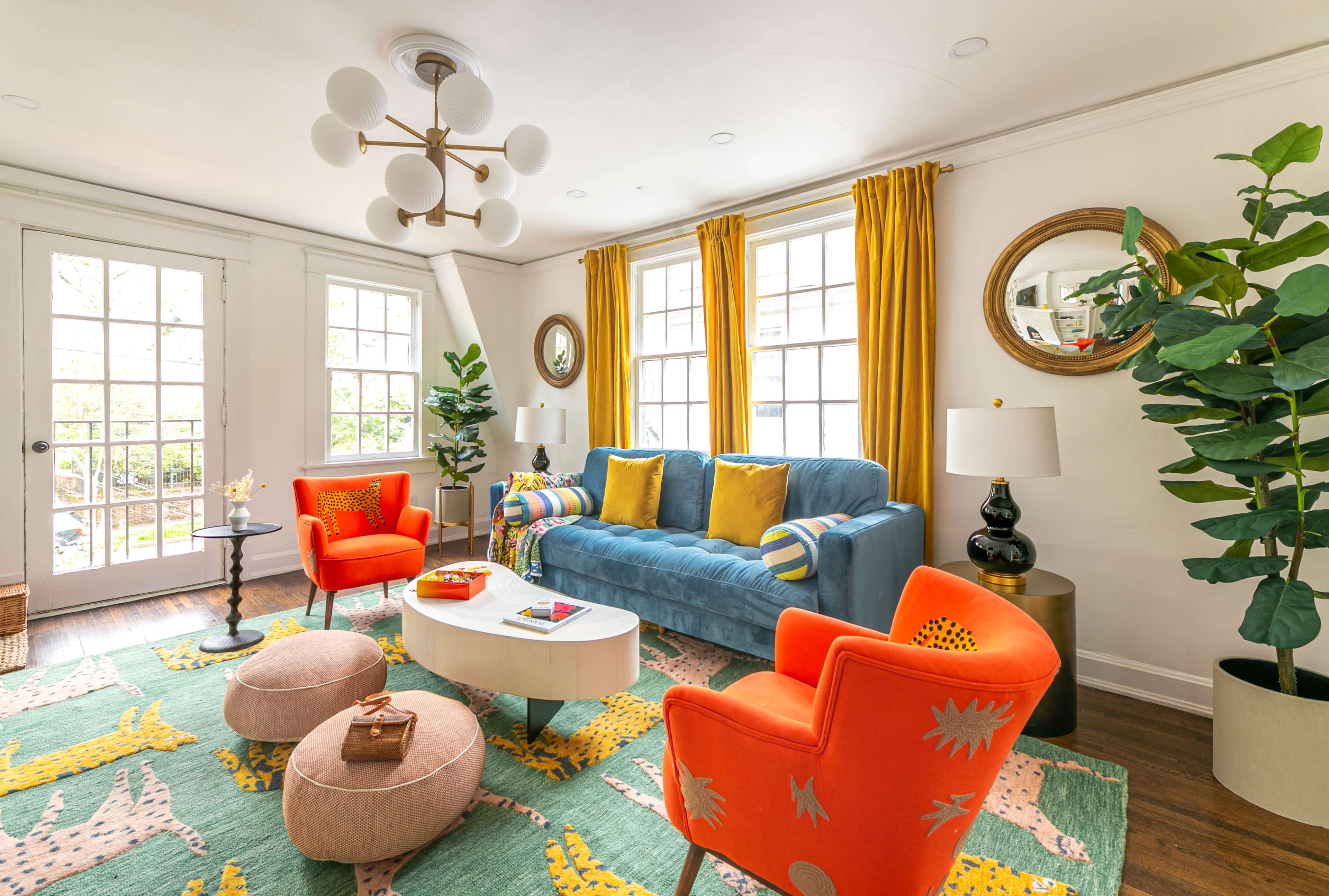 A brightly decorated living room features a blue sofa, orange chairs, a white coffee table, and large windows with yellow curtains.