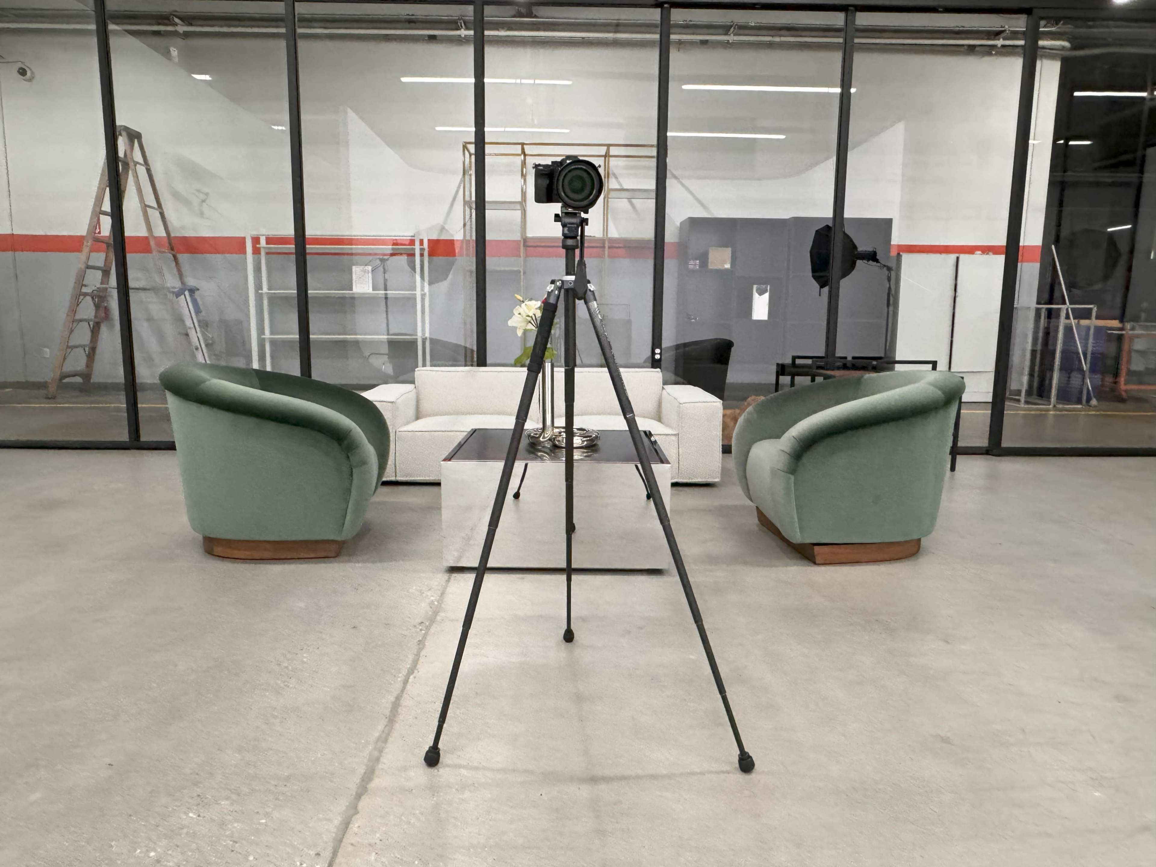 A camera on a tripod is positioned facing a seating area with two green chairs and a white sofa, all set within a spacious industrial room.
