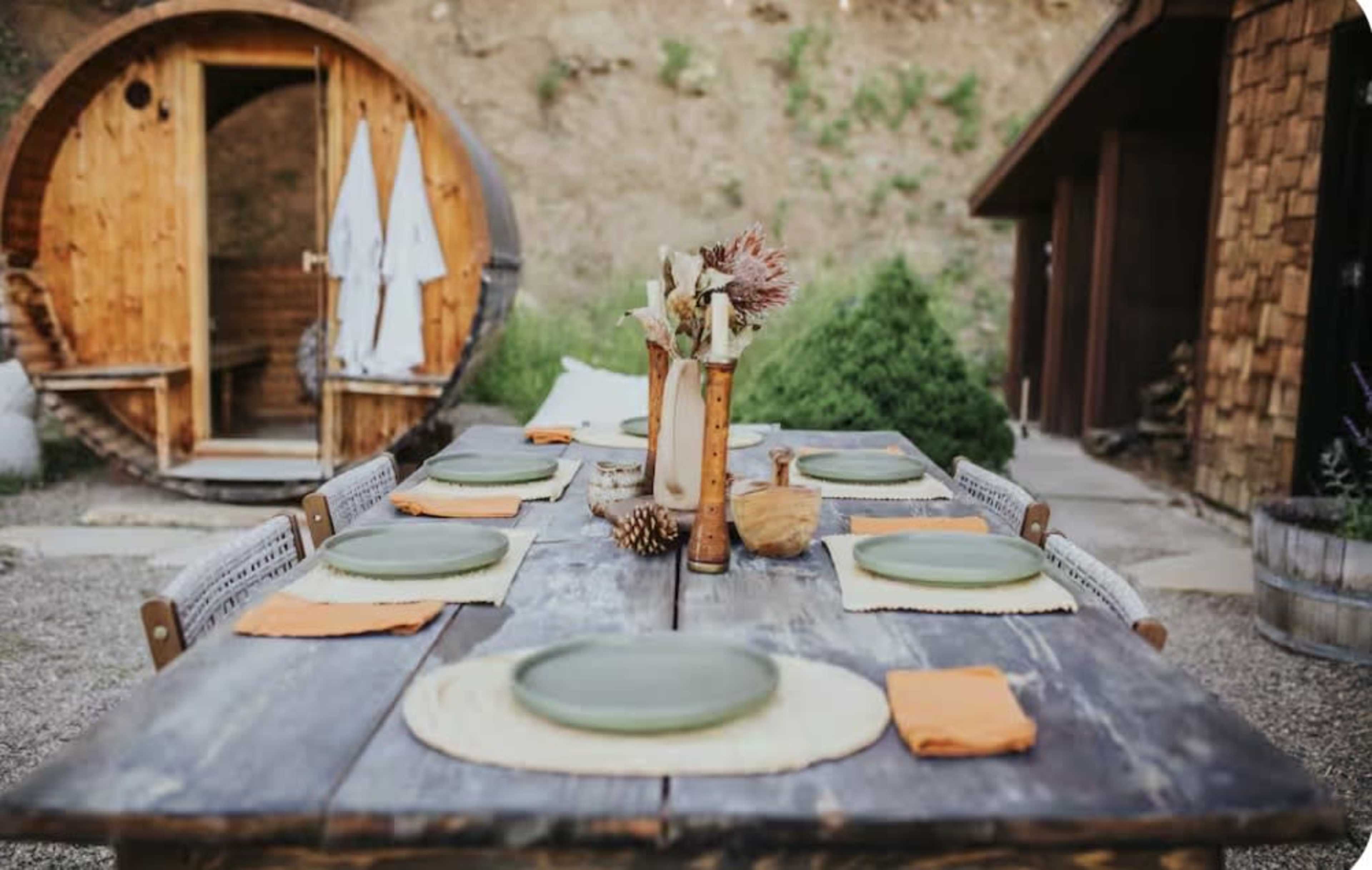 A rustic outdoor dining setup features a wooden table with green plates and orange napkins, complemented by a nearby wooden structure resembling a sauna.