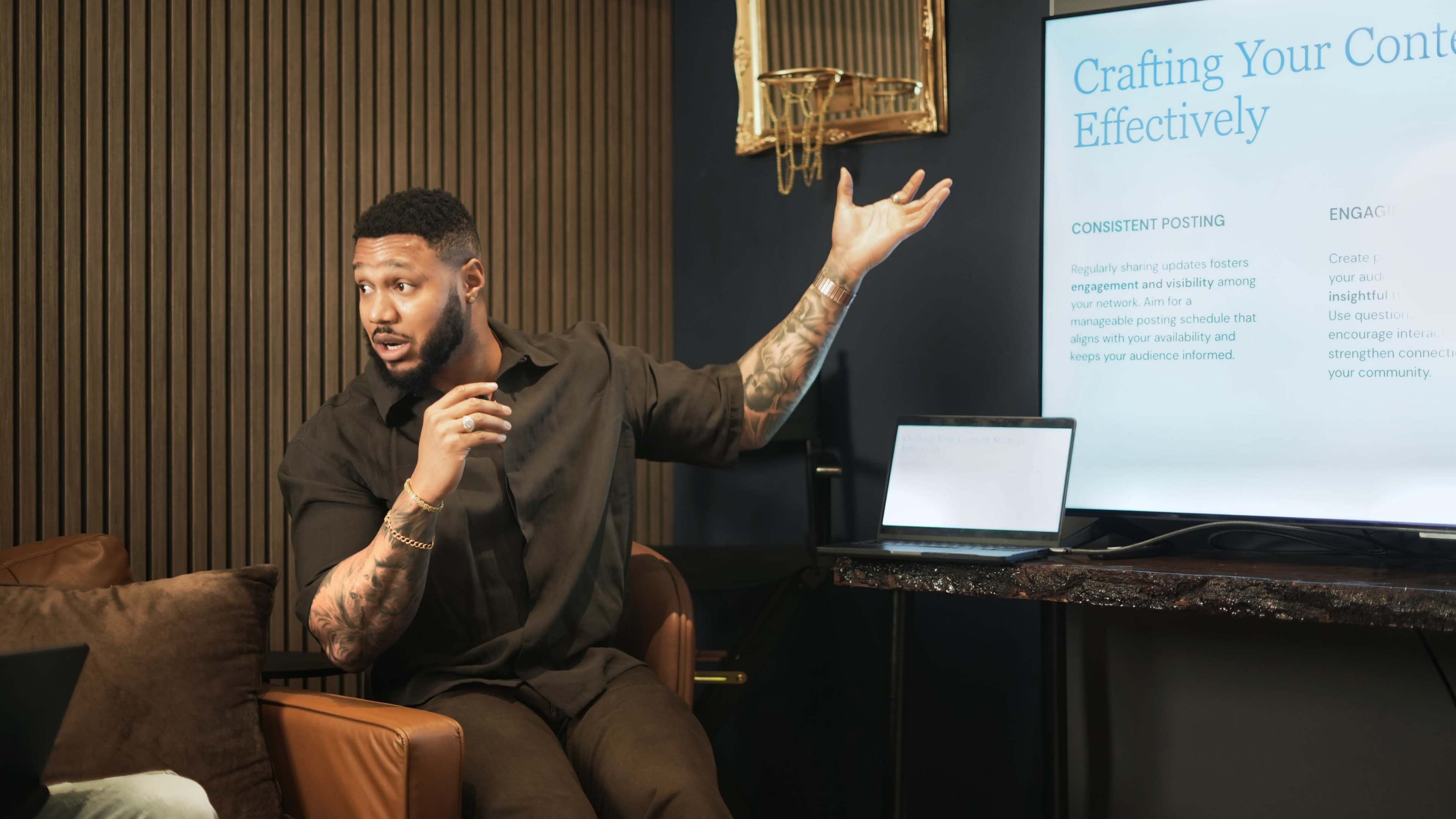 A man with tattoos gestures emphatically while presenting information on a screen during a meeting.