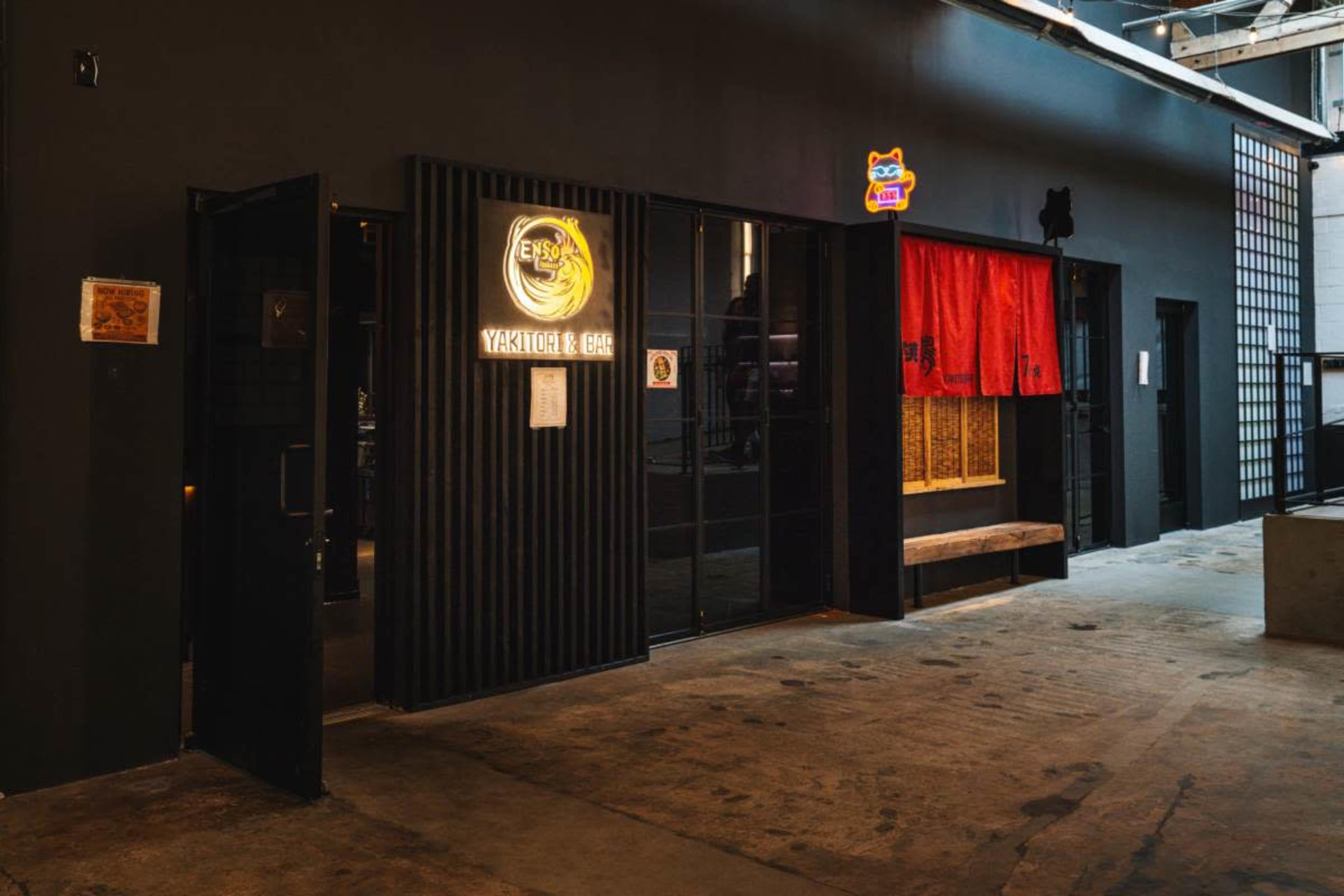The image shows the entrance of a restaurant with a black exterior, featuring a lit sign that reads "YAKITORI & BBQ," along with a red curtain and a wooden bench outside.