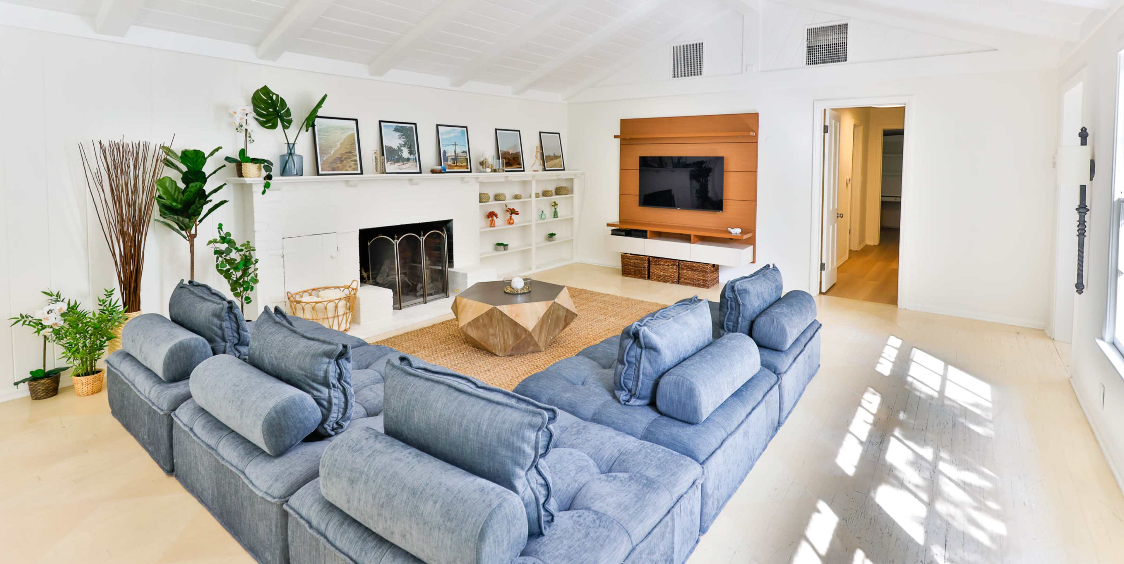 The living room features a large sectional sofa facing a wall-mounted television, with a fireplace and various plants surrounding a central coffee table.