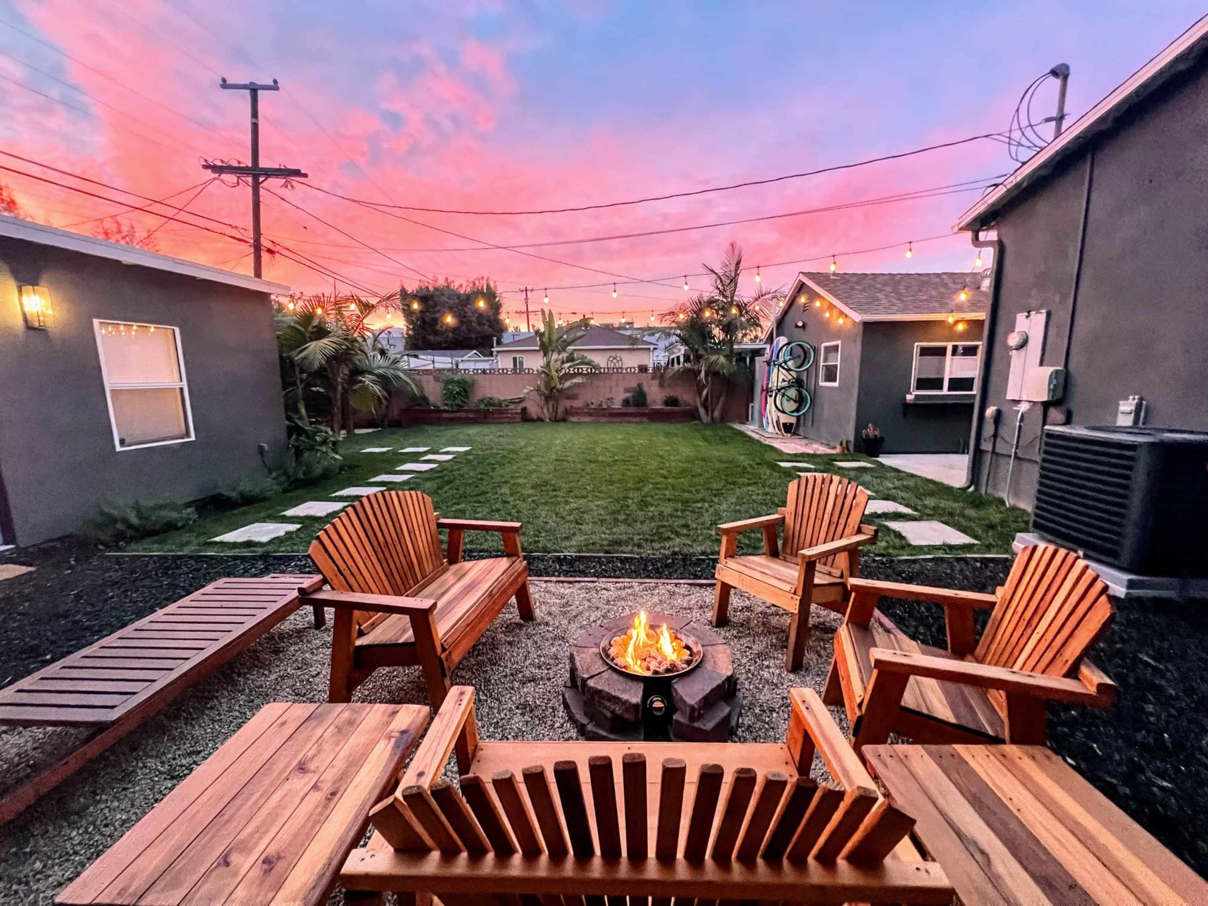 A cozy outdoor space features four wooden chairs surrounding a fire pit, with a well-maintained lawn and colorful sunset skies in the background.