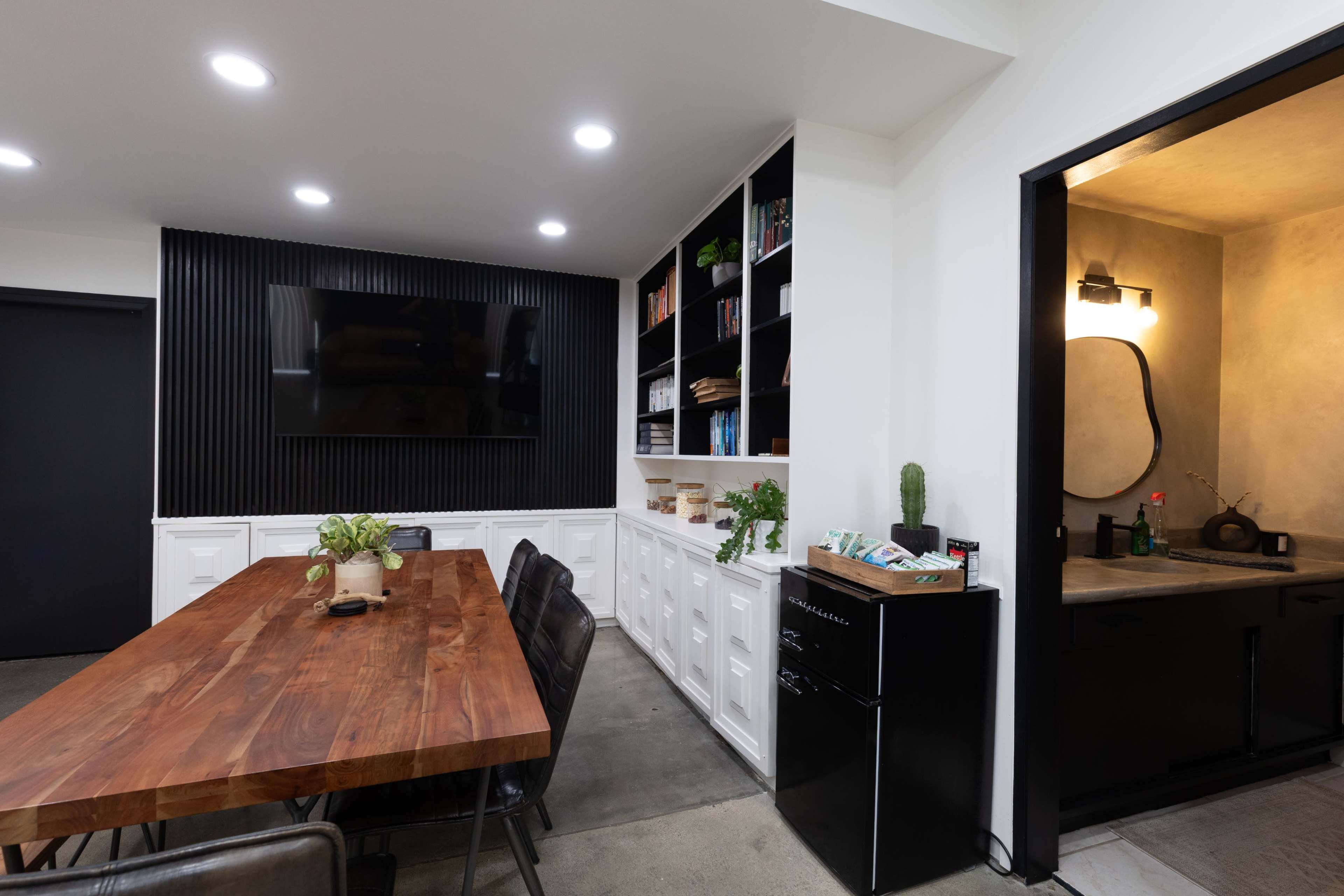 The image shows a modern dining area with a large wooden table, black chairs, a wall-mounted television, built-in bookshelves, and a small kitchen area with a fridge and sink.