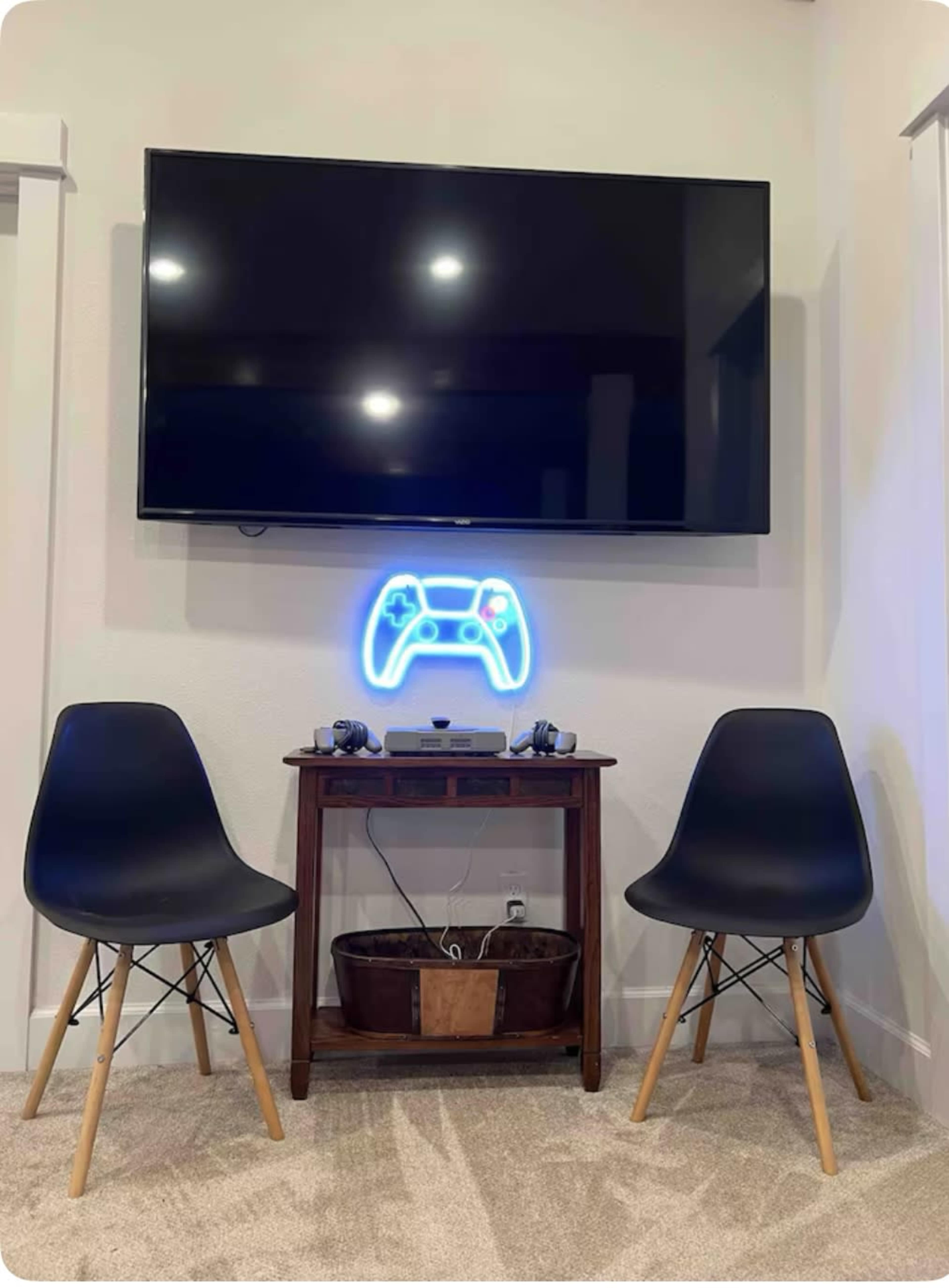 A large television is mounted on a wall above a wooden table with gaming consoles and controllers, flanked by two black chairs.