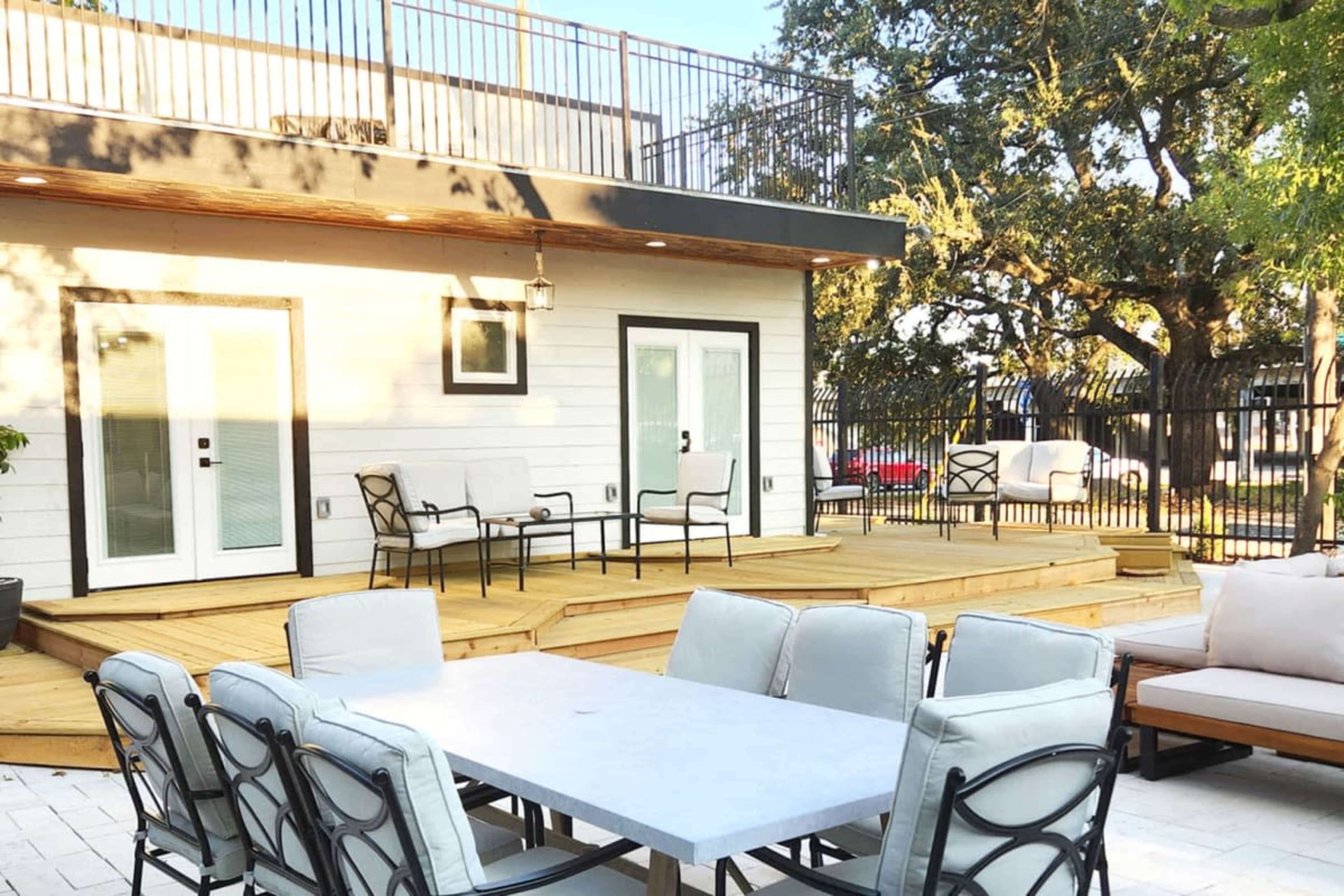 A modern outdoor patio area with a wooden deck, a large dining table, and seating arrangements near a building.