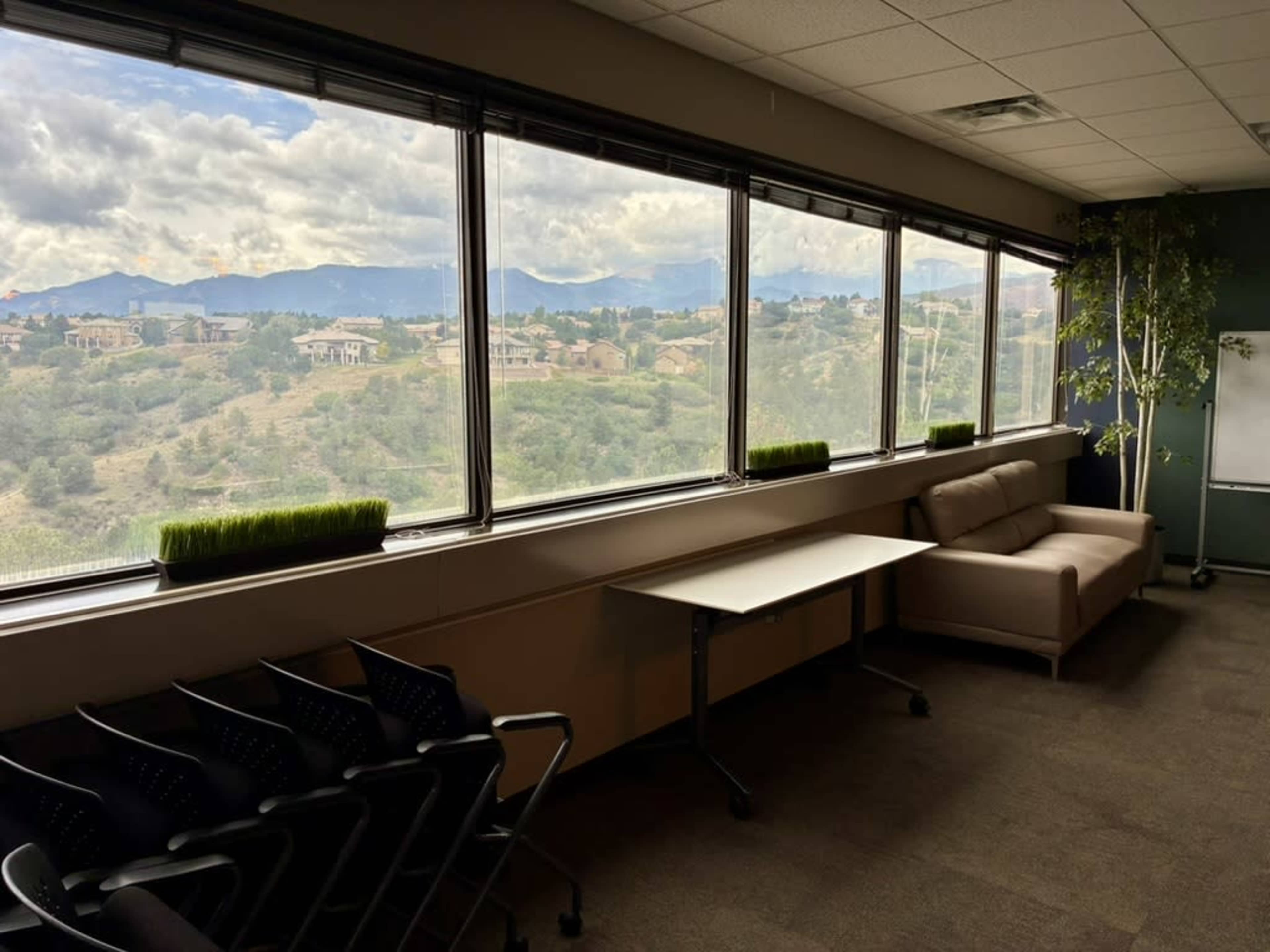 The image shows a spacious office area with large windows overlooking rolling hills and mountains, furnished with a couch and a table.