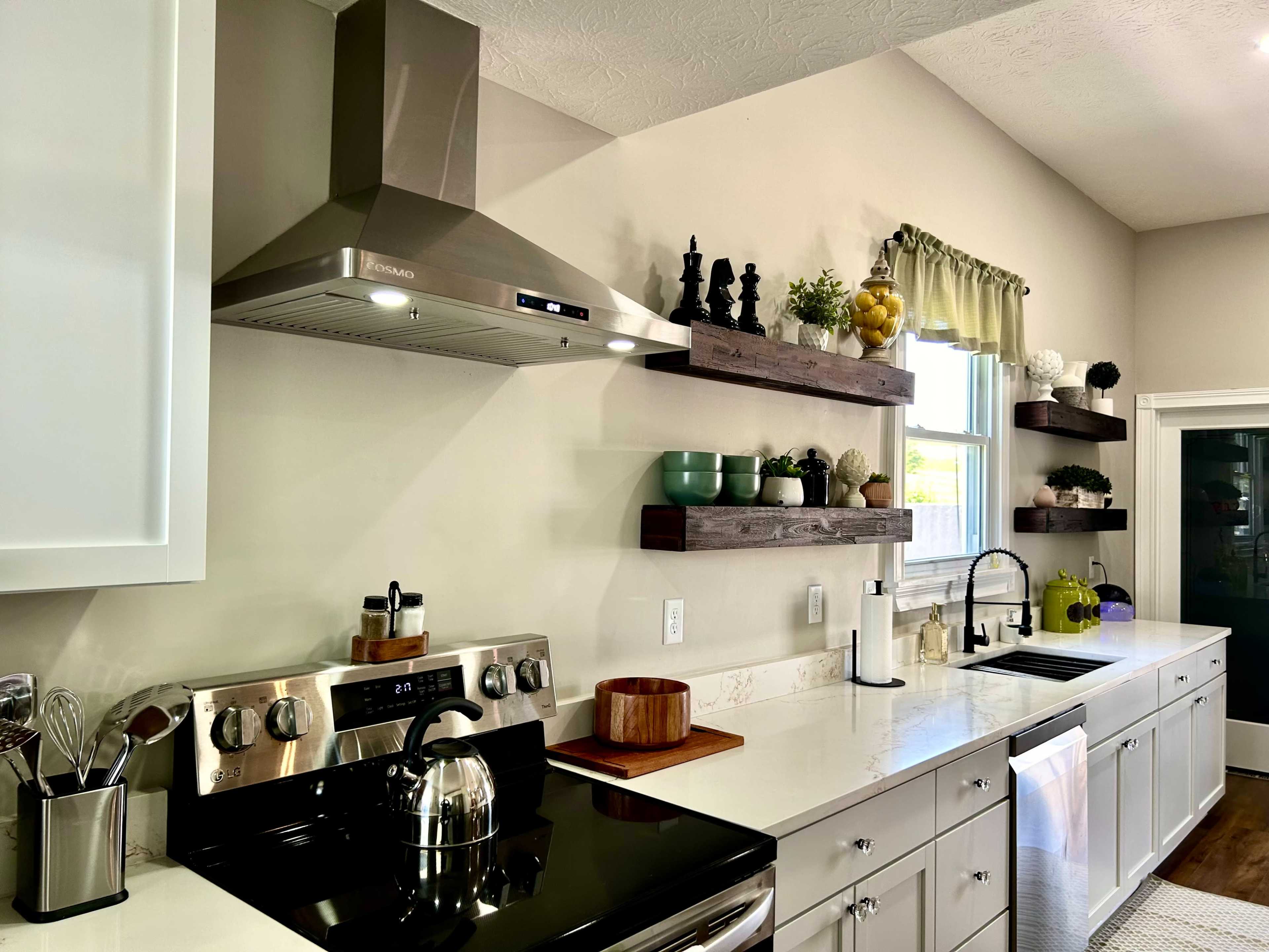 The image shows a modern kitchen with a stainless steel range hood, a black stove, white cabinets, and floating wooden shelves displaying various kitchen items and plants.