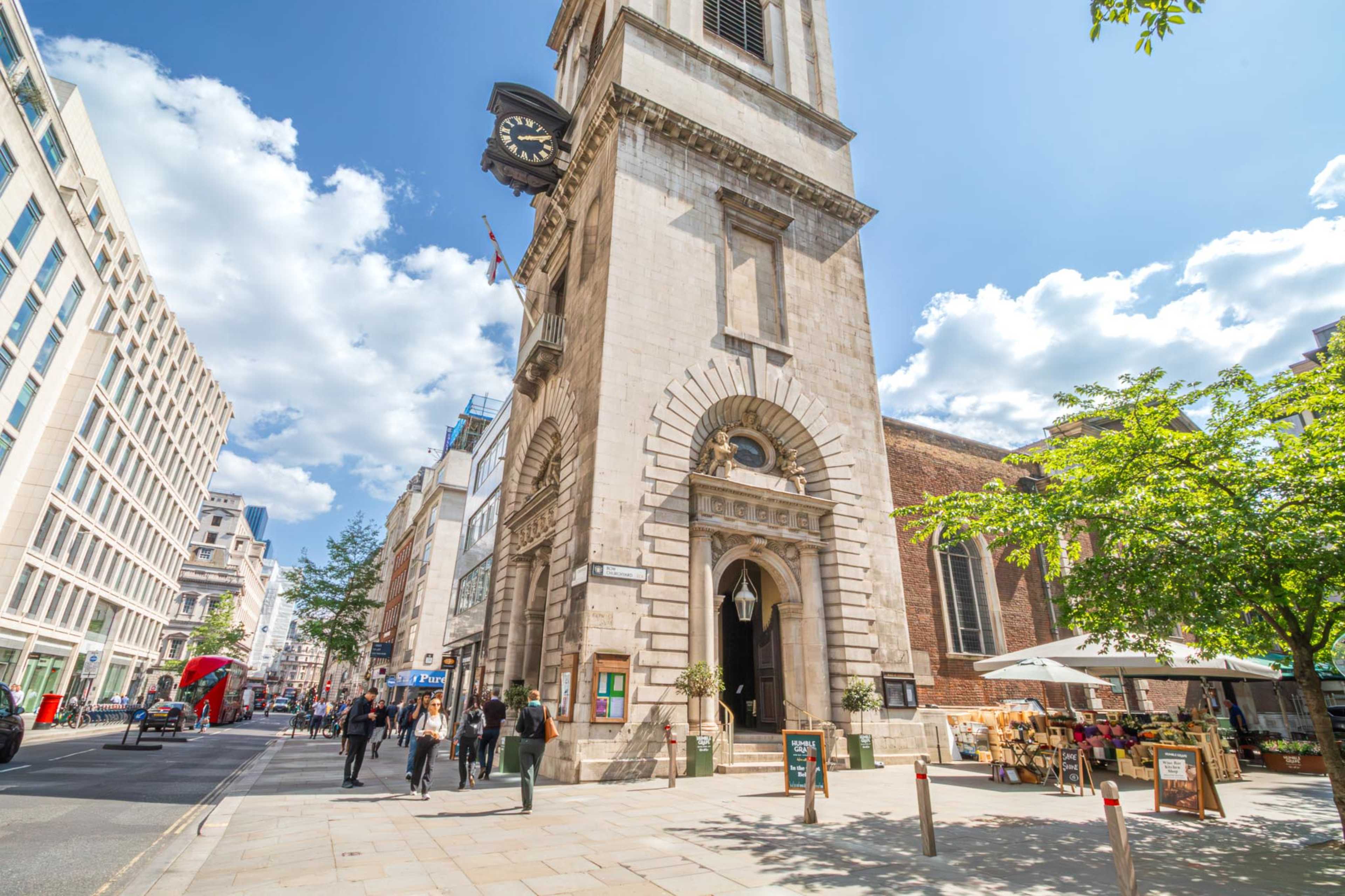 A historic building with a clock tower stands at the corner of a busy street, surrounded by modern architecture and pedestrians.