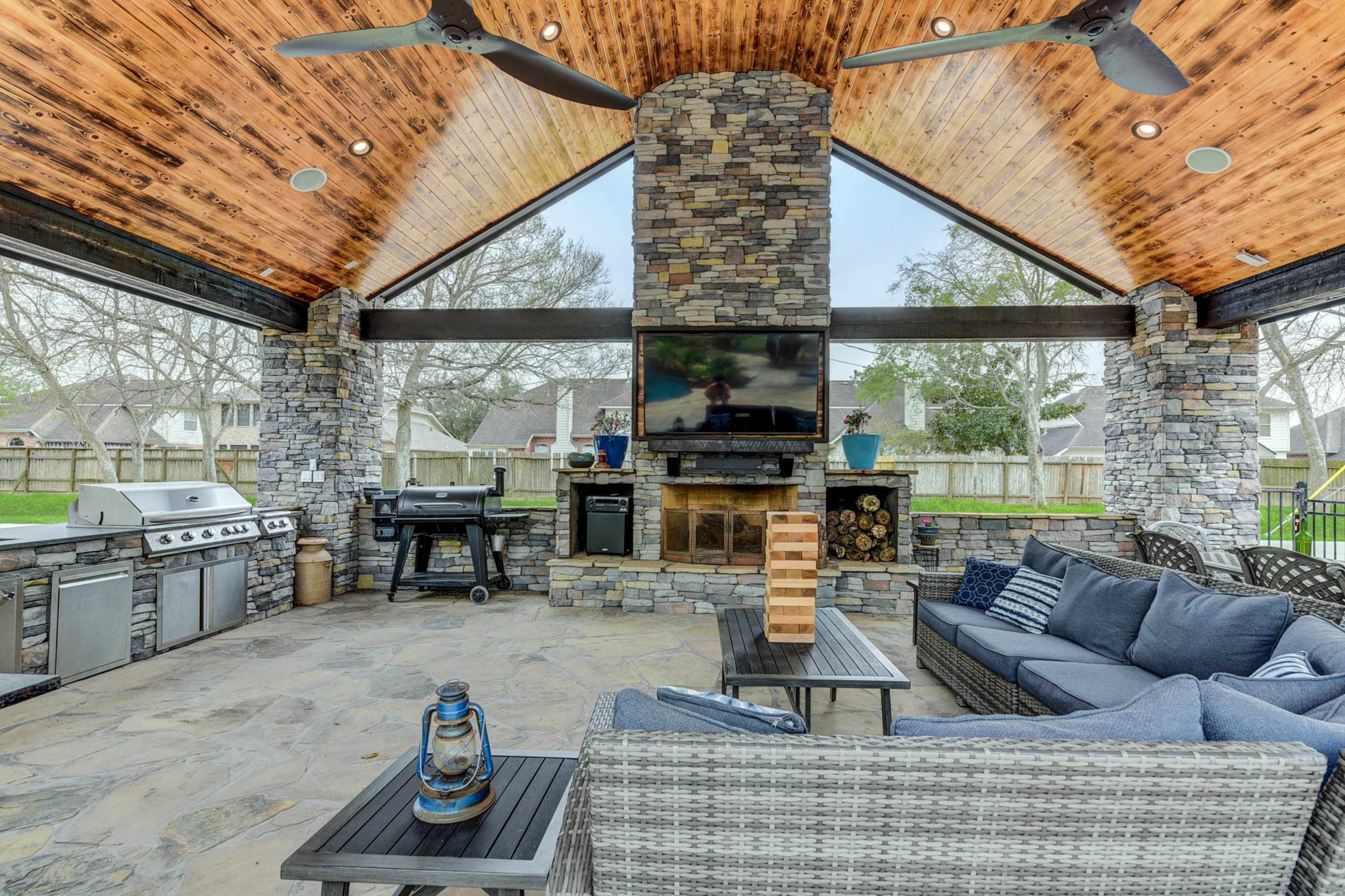 An outdoor patio with stone walls, a wooden ceiling, a grilling area, a seating arrangement, and a mounted television.