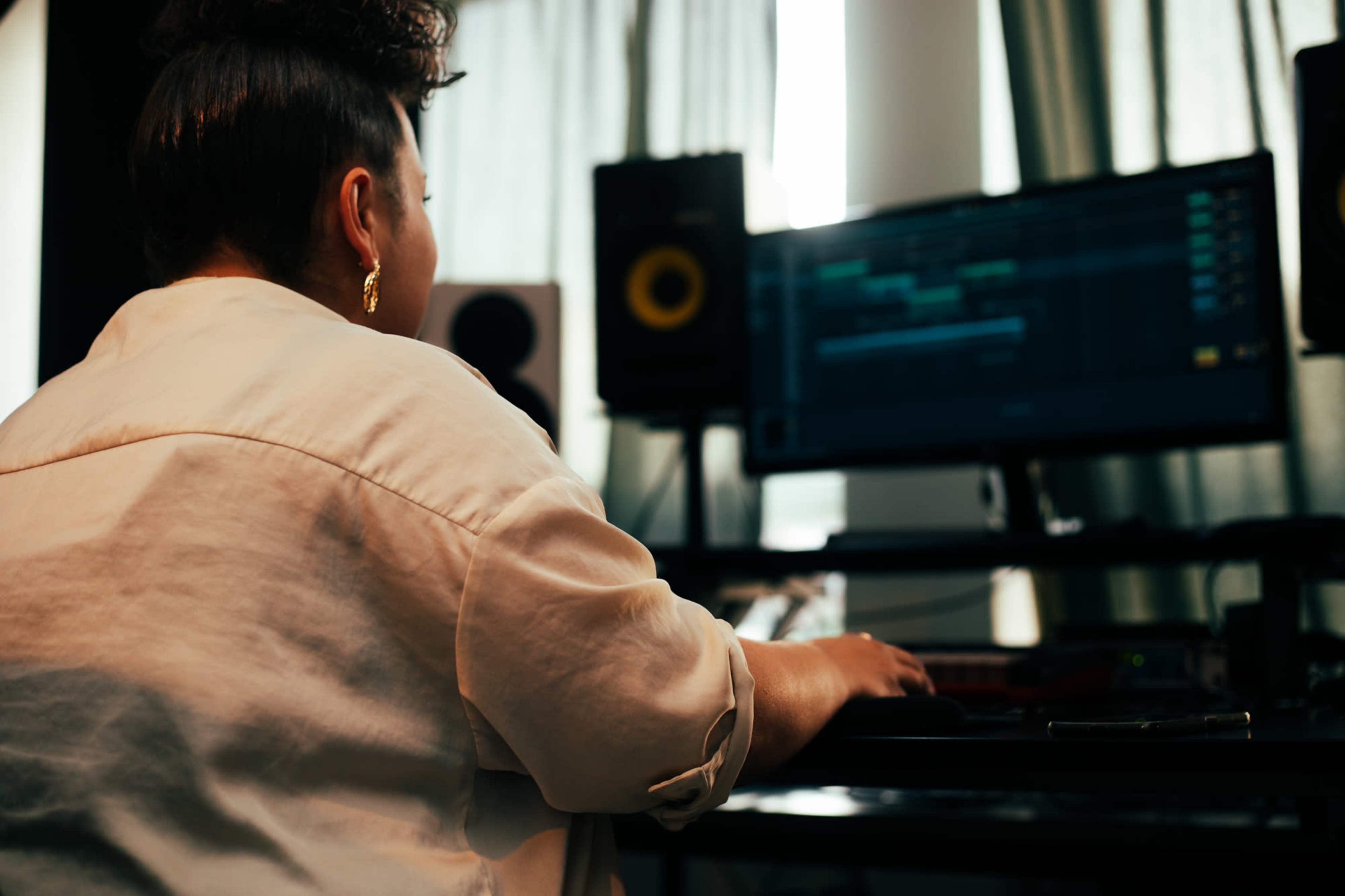 A person with curly hair sits at a desk working on a computer in a music production studio.